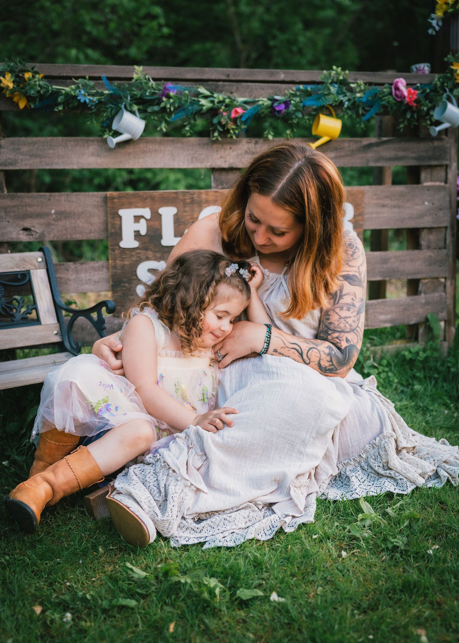 A tattooed mother tenderly embraces her young daughter in a boho outdoor setting with floral decorations.