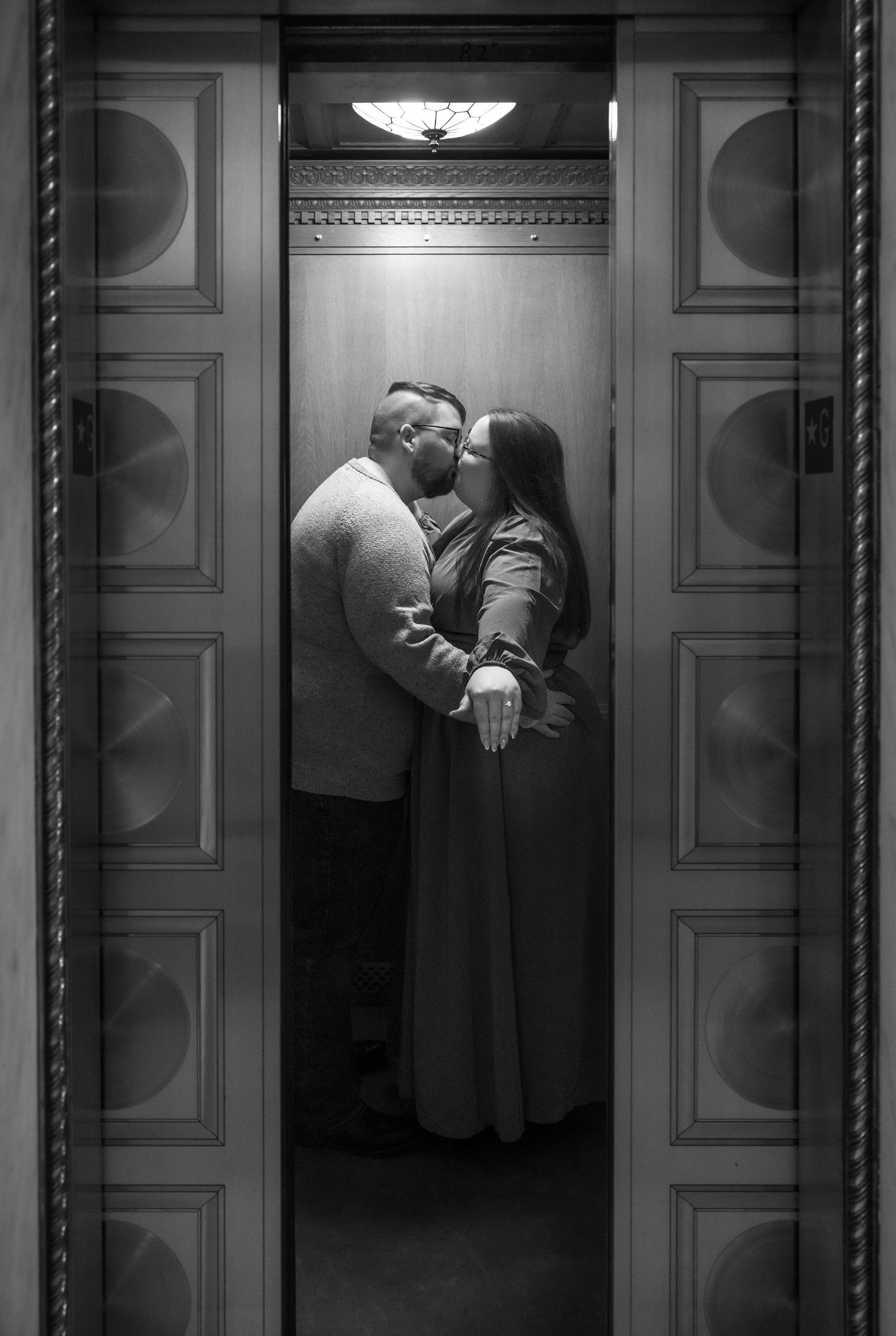 Black and white photo of couple kissing inside an ornate wooden elevator.