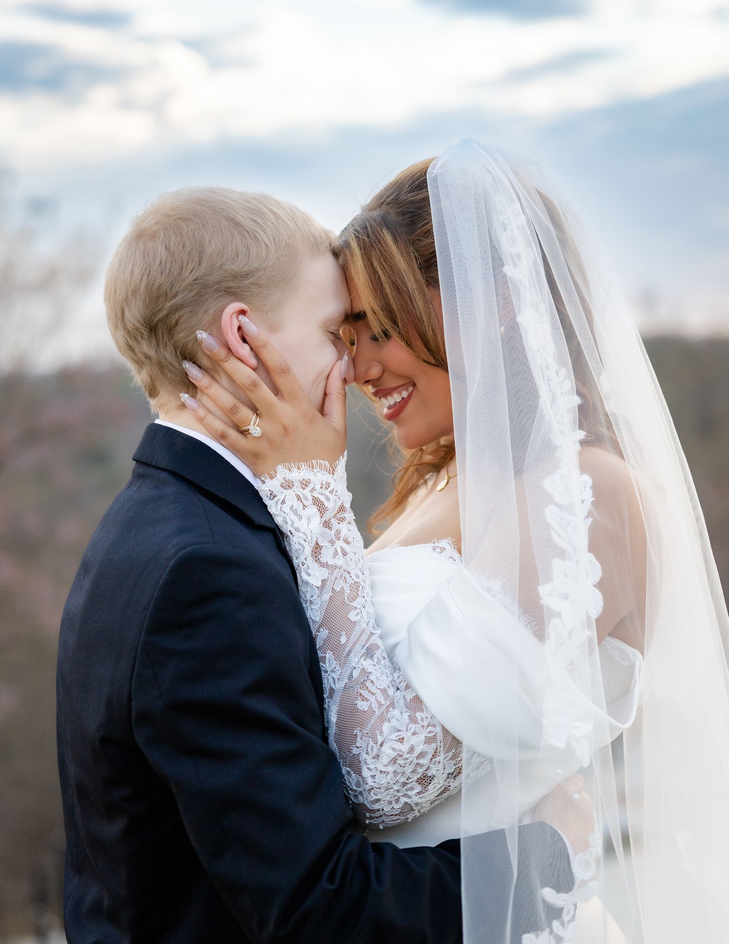 Joyful newlyweds touch foreheads tenderly under bridal veil against dramatic cloudy sky.