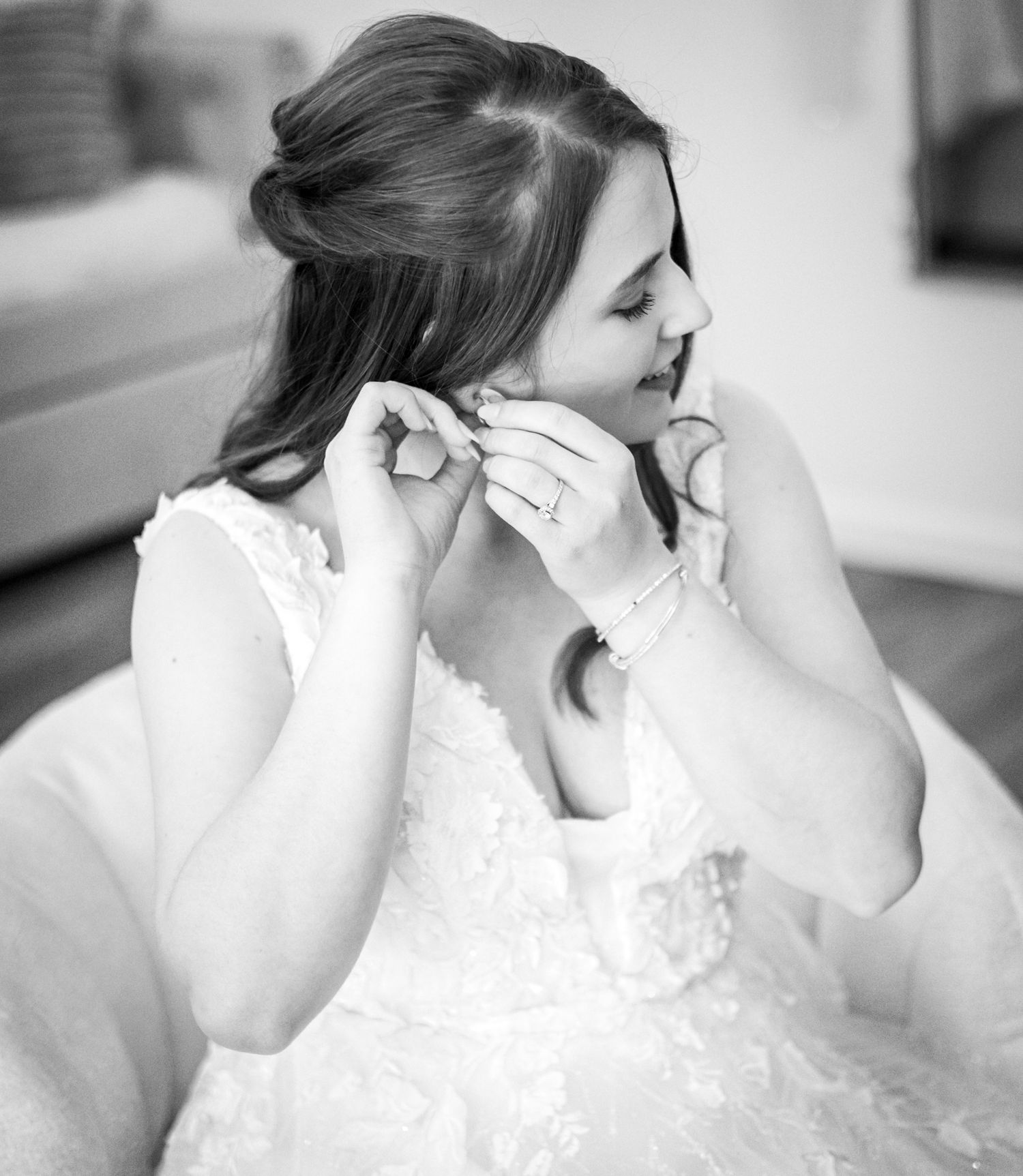 Black and white photo of a bride in lace wedding dress putting on earrings while getting ready.