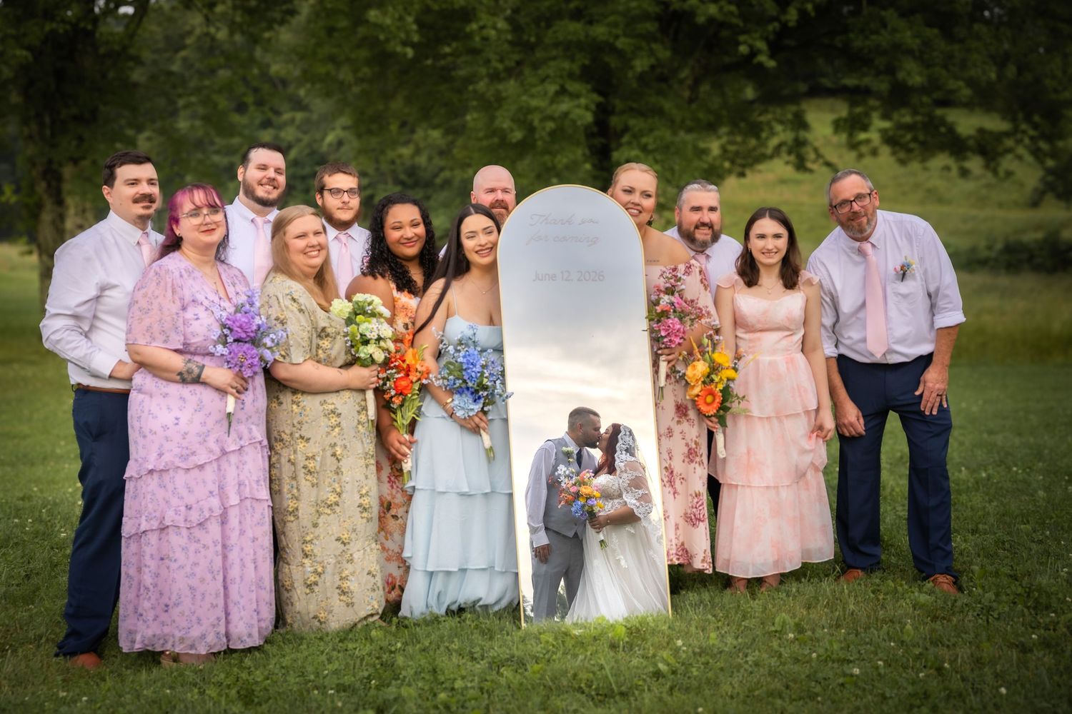 Wedding party poses outdoors on green lawn with bride and groom kissing beside tall mirror.
