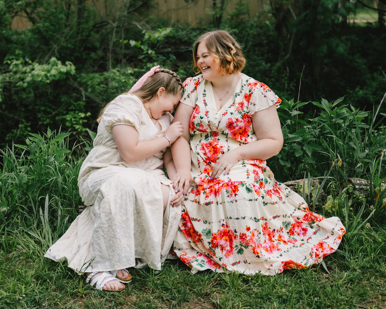Two women in floral and cream dresses laughing together outdoors in a lush green garden setting.