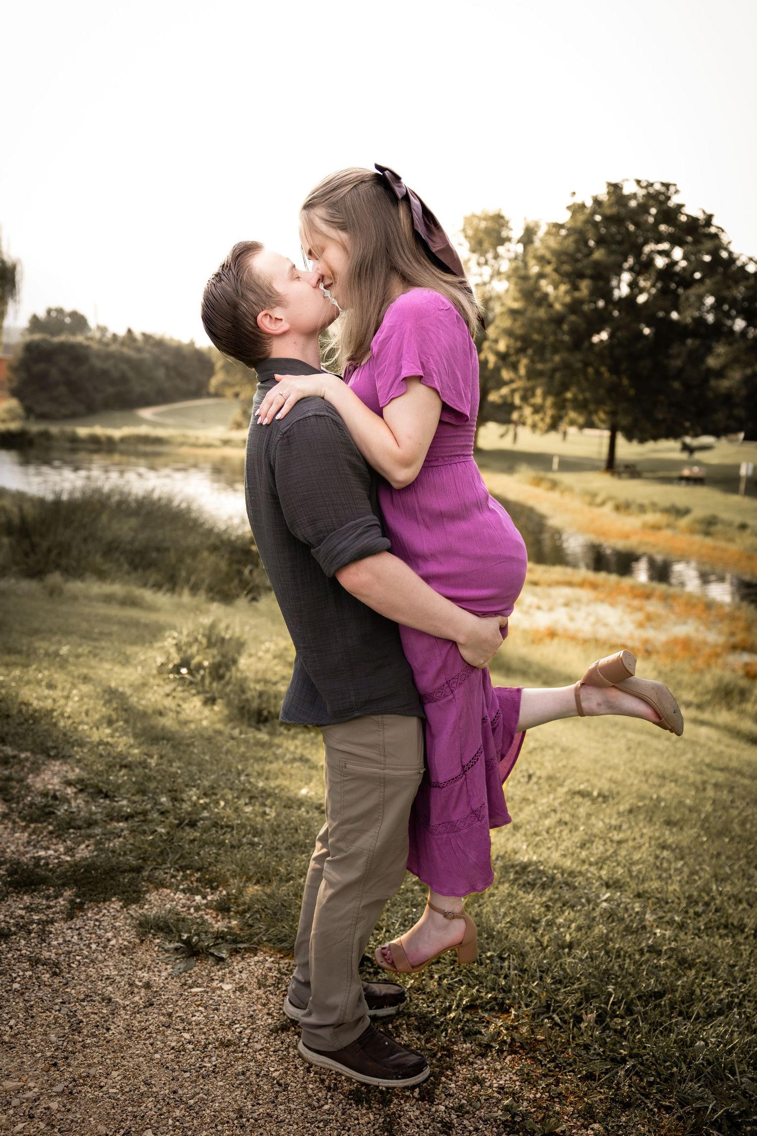 A couple shares a romantic kiss outdoors, the man lifting the woman in a pink dress near a scenic pond.