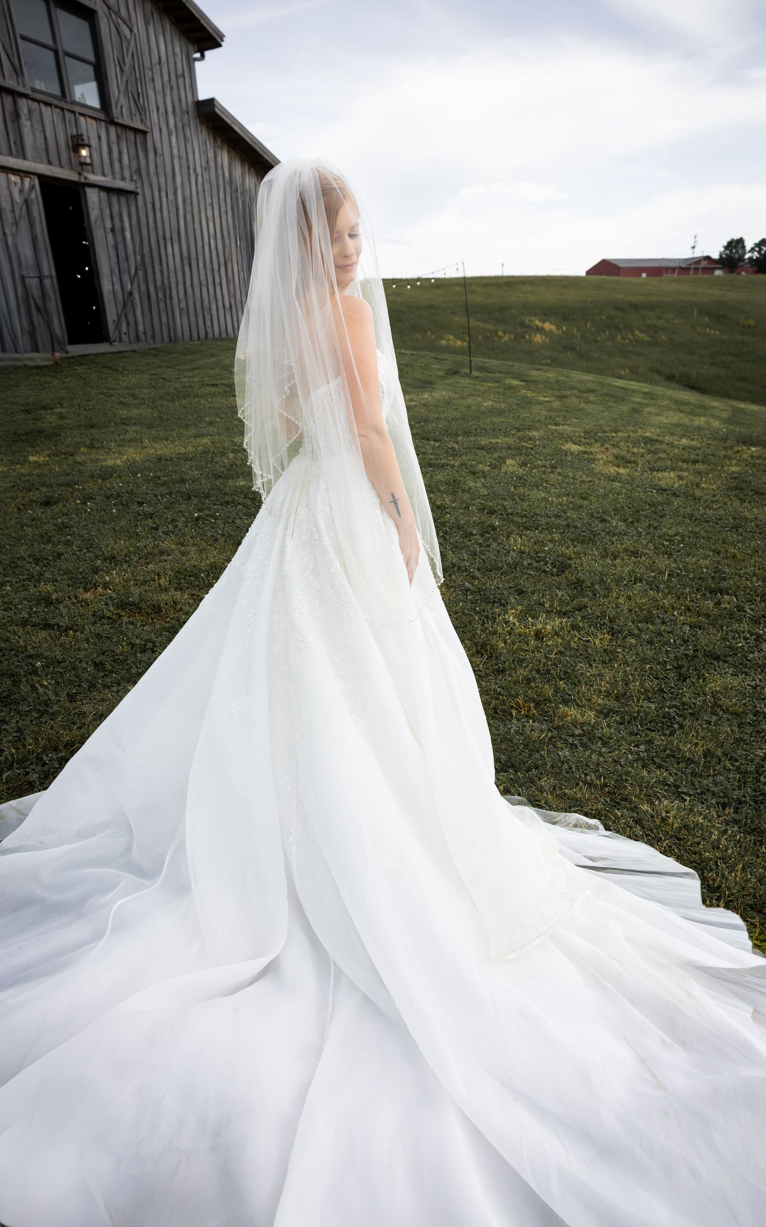 Bride in elegant white wedding gown with long train and veil stands outdoors near rustic barn on green hillside.
