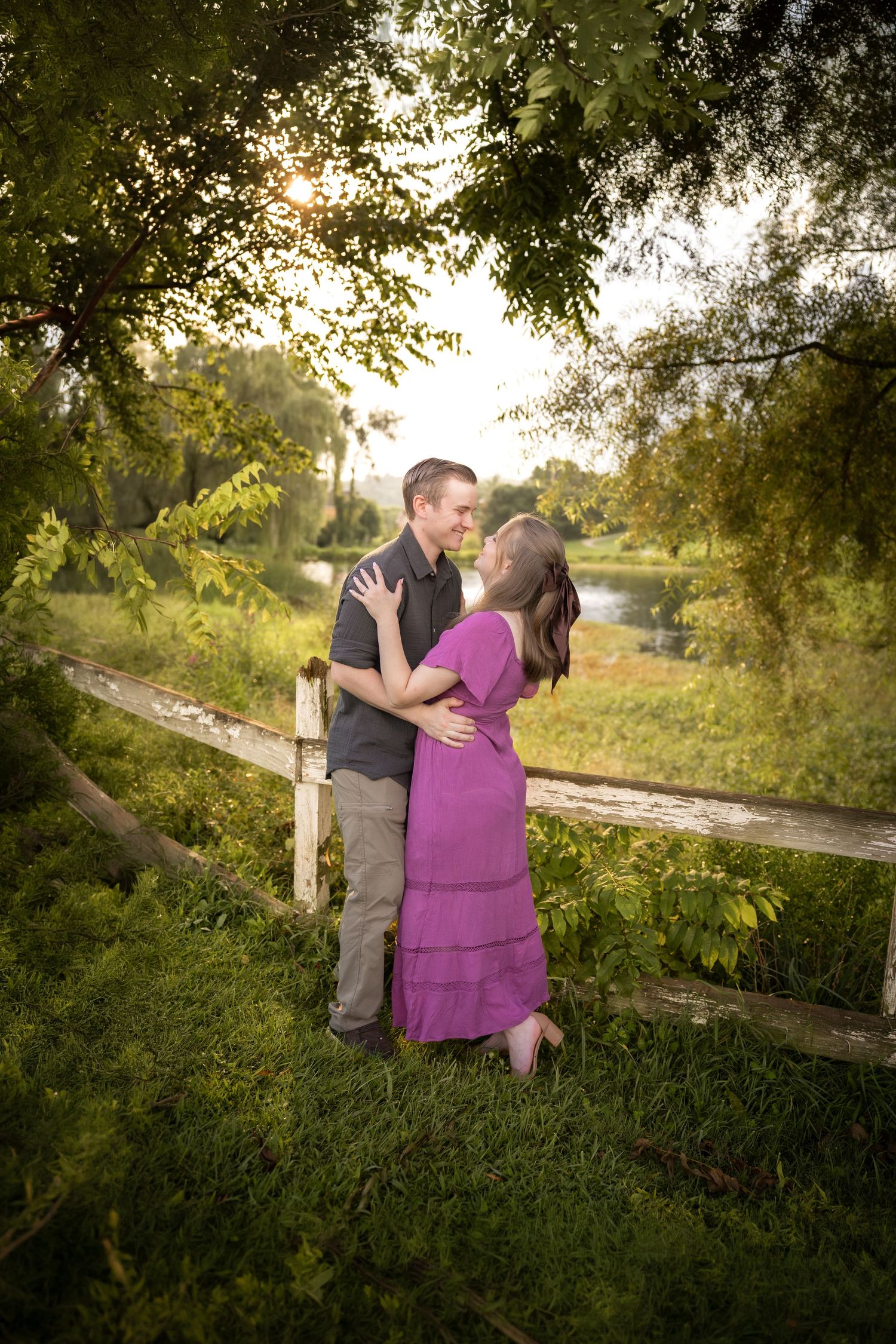 Couple sharing a romantic moment near a rustic fence surrounded by lush green trees and soft golden sunset light.