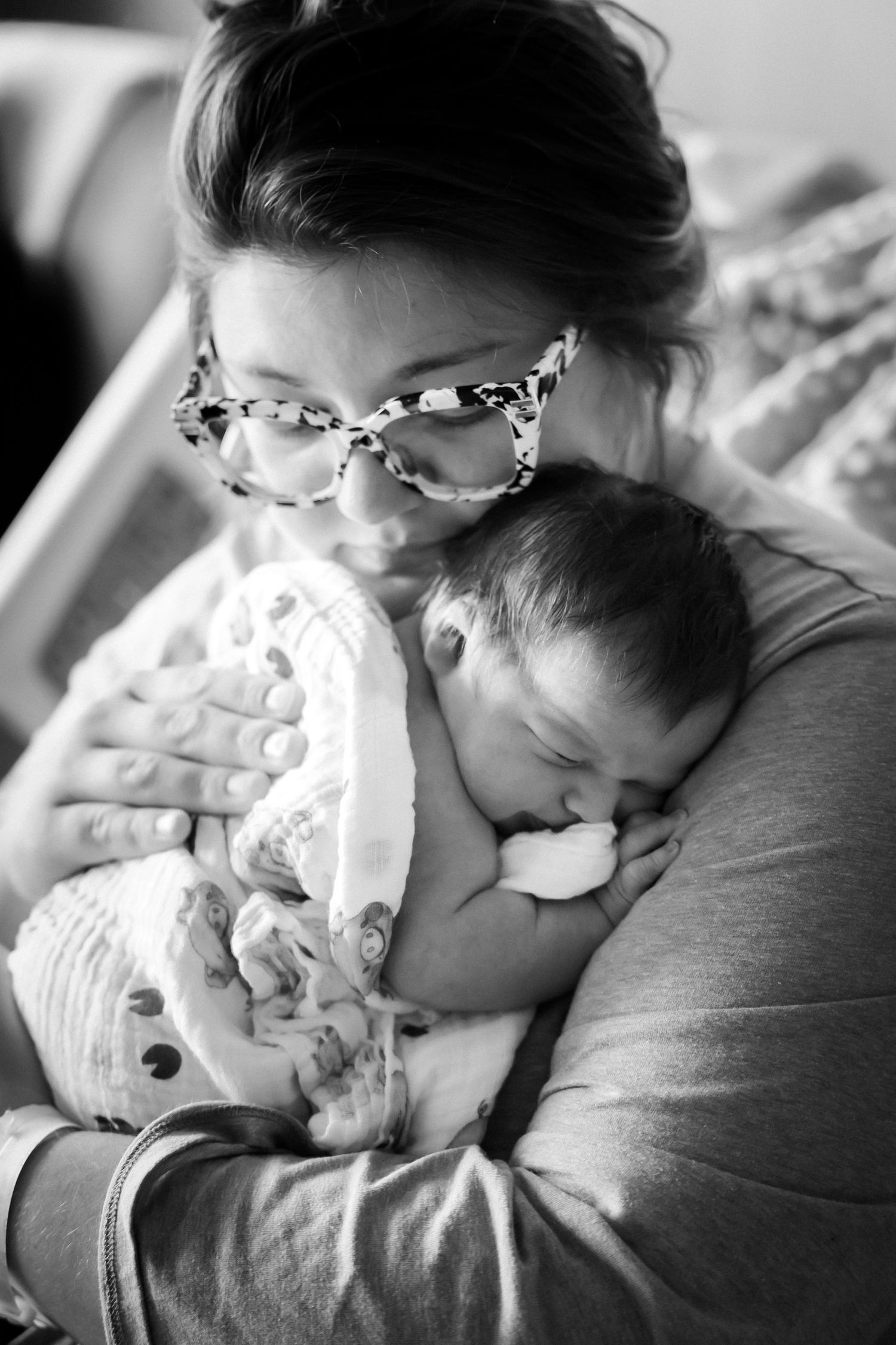 Mother with glasses tenderly holding and kissing her sleeping newborn baby in black and white.