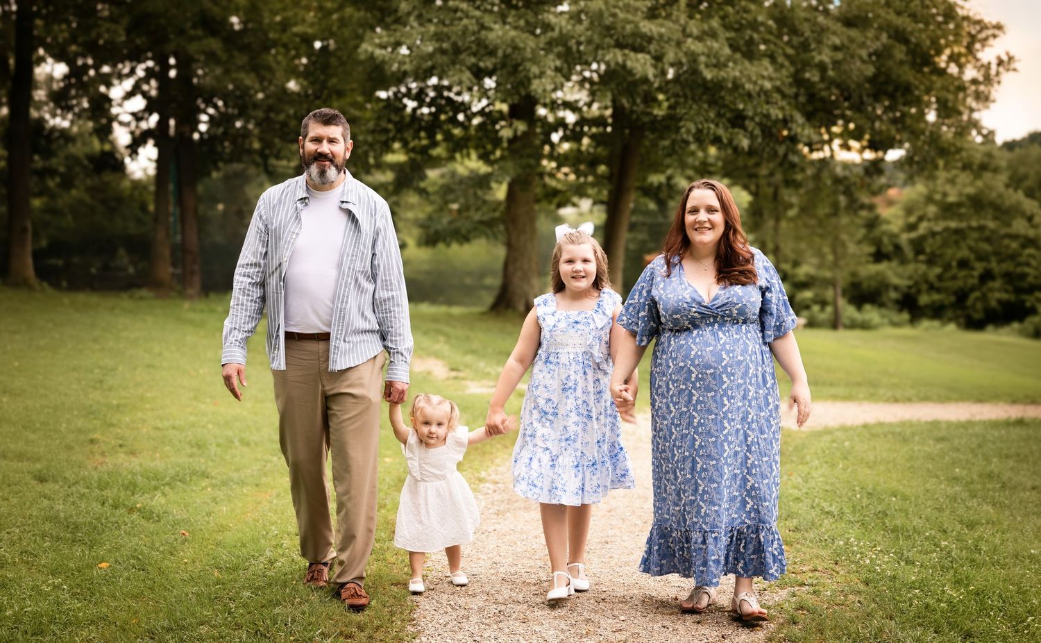 Family of four walks hand-in-hand on a garden path, parents with two young daughters in floral dresses outdoors.