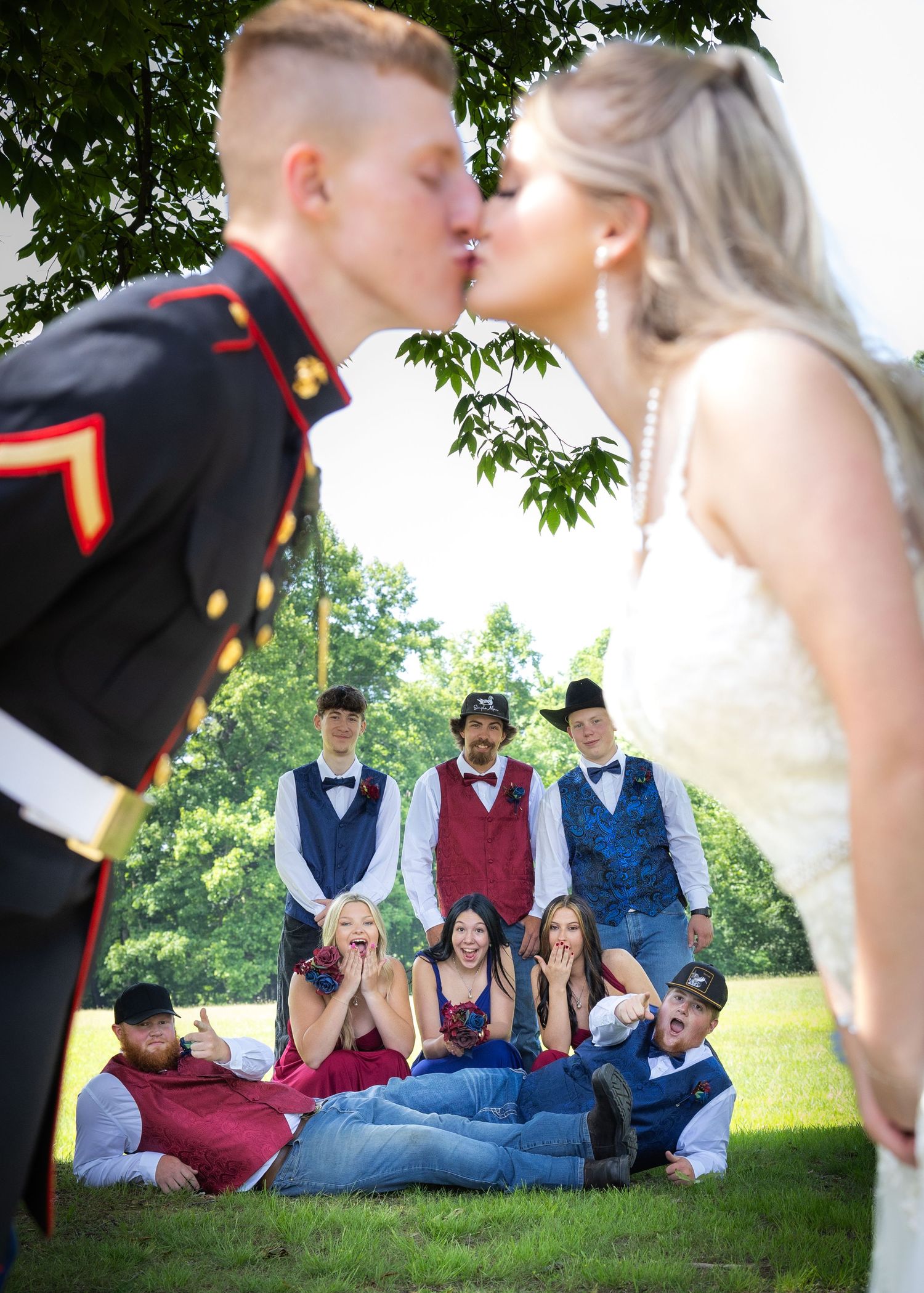 Marine groom kisses bride at outdoor wedding while bridal party reacts with joy and surprise behind them.