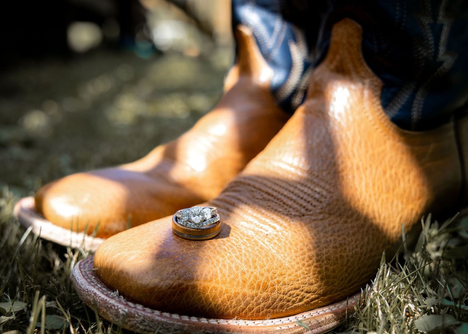 Close-up of tan leather cowboy boots with a diamond ring resting on top, set against a rustic outdoor background.