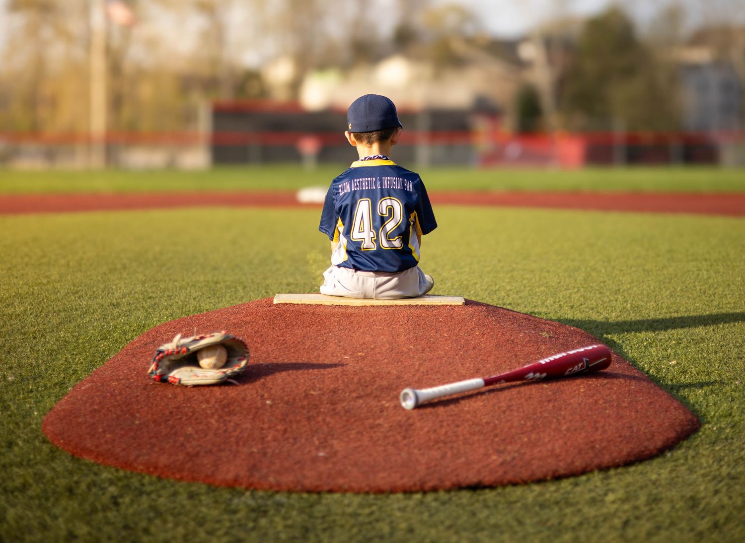 Child in baseball uniform number 42 sitting alone on pitching mound with bat and glove.