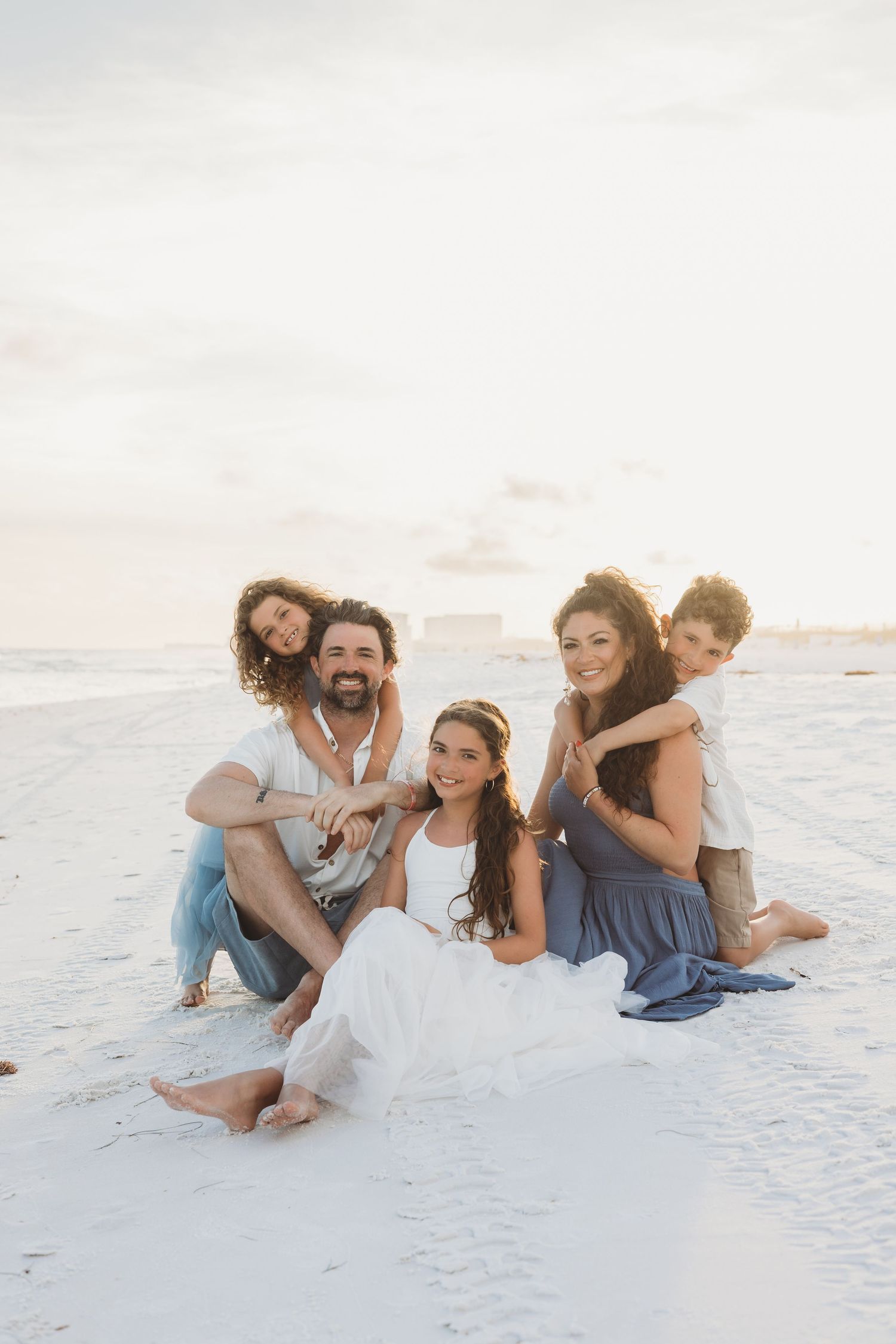 Happy family of five posing together on a white sand beach at sunset, smiling at the camera.
