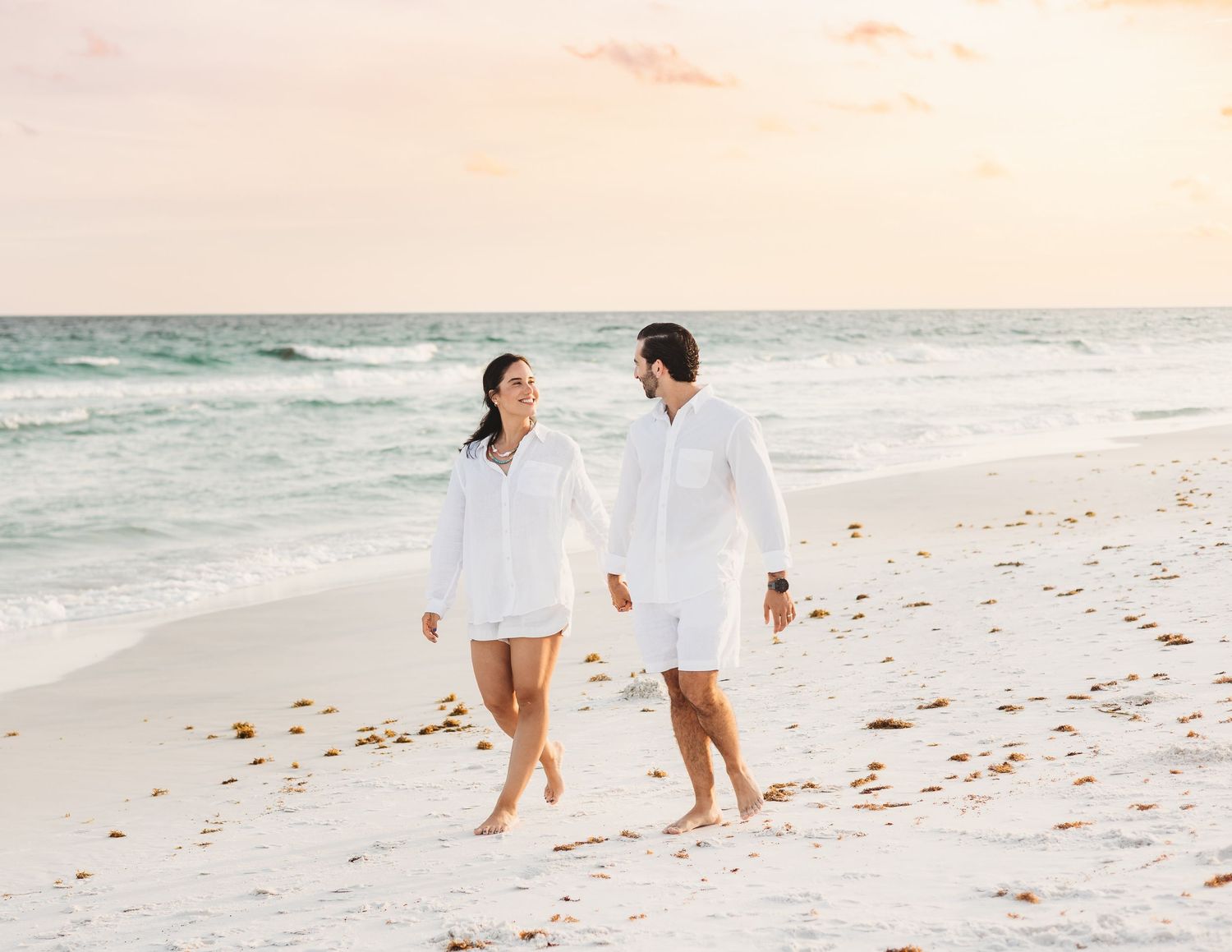 Couple in matching white outfits walking hand in hand on a sandy beach at sunset.