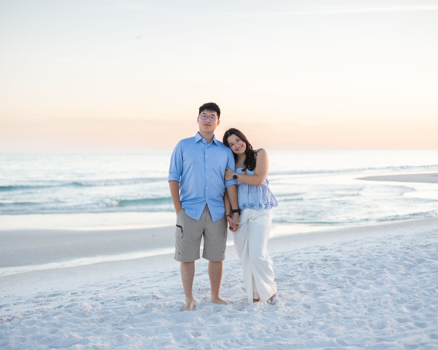 A couple poses barefoot on a sandy beach at sunset, the woman leaning on the man in a blue shirt and khaki shorts.