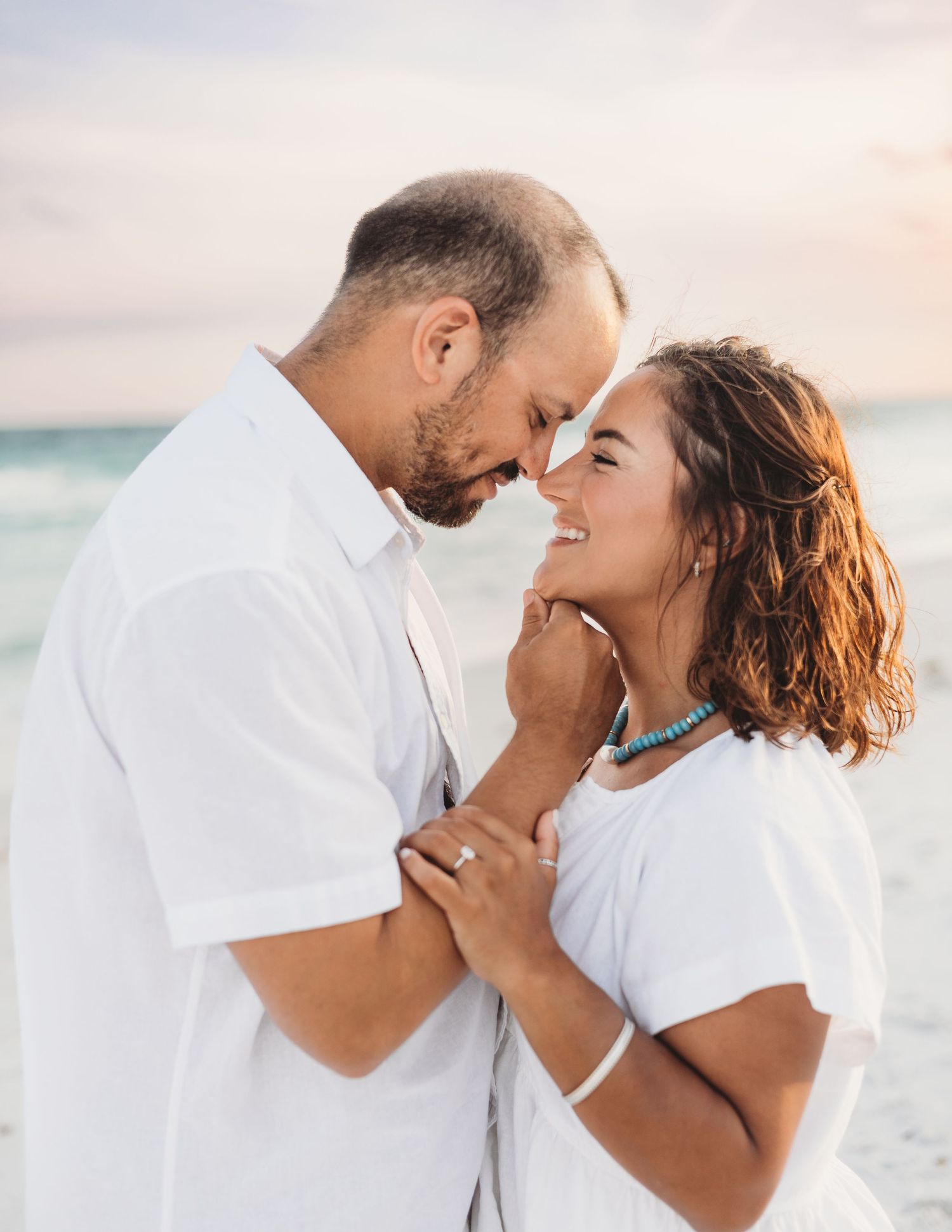 Romantic couple in white outfits touching foreheads and smiling on a beach at sunset.