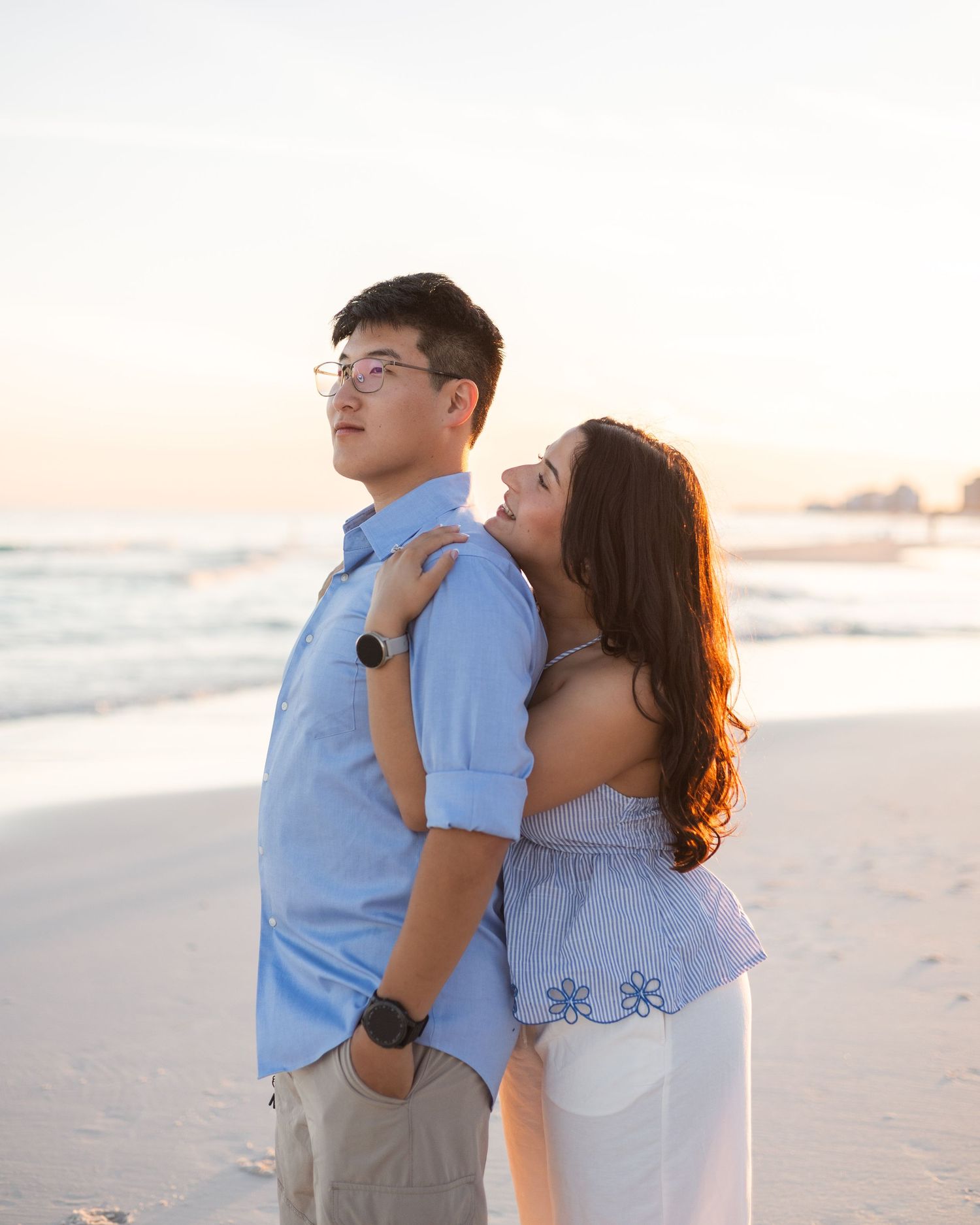 Couple posing on a beach at sunset, woman embracing man from behind, both wearing light blue outfits.