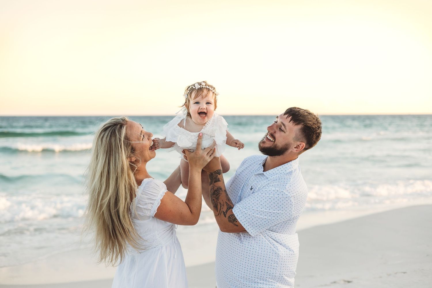 Happy family with tattooed dad lifting laughing baby girl on a sunny beach, mom smiling in white dress.