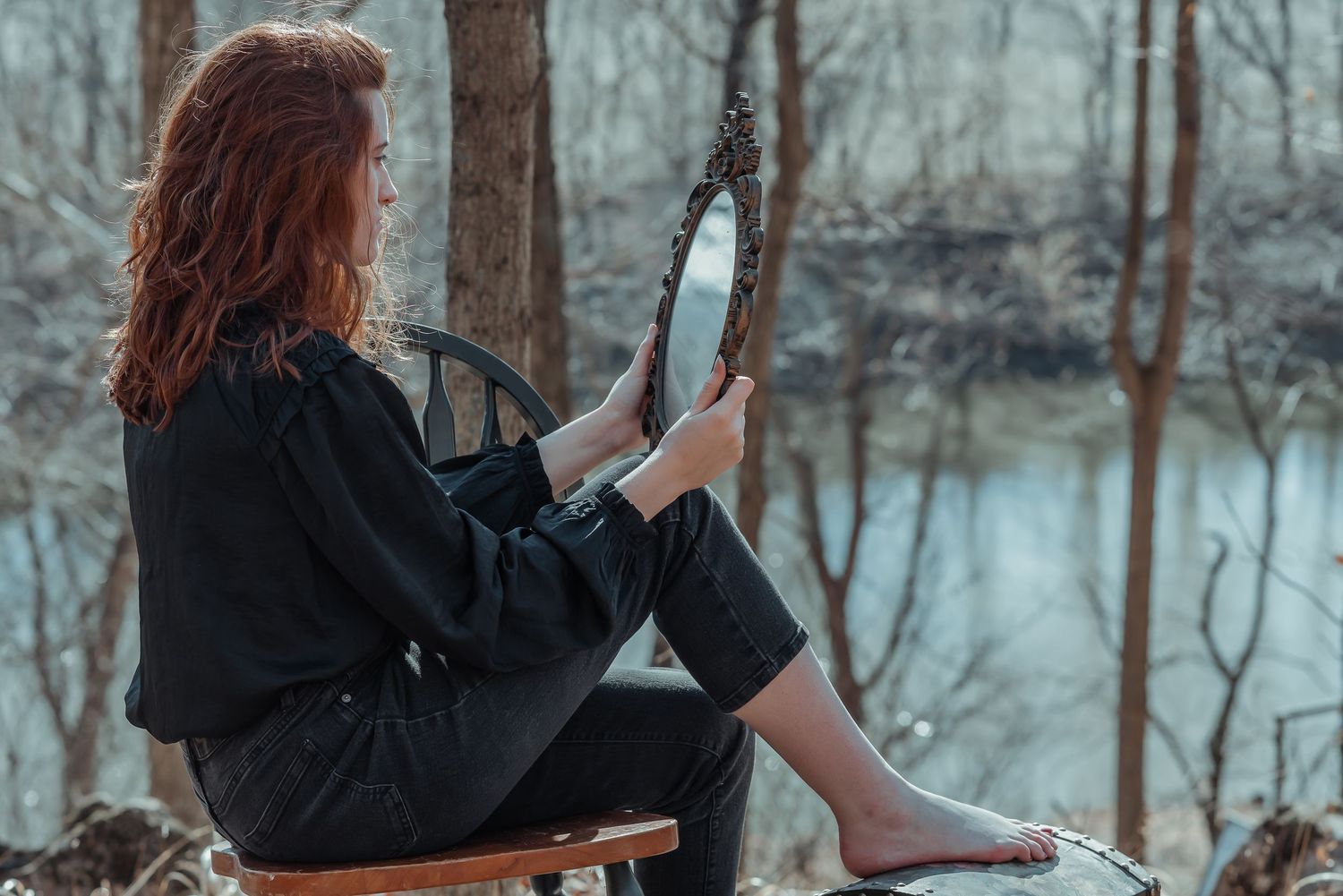 A red-haired woman in black sits on a chair holding an ornate mirror in a forest setting.