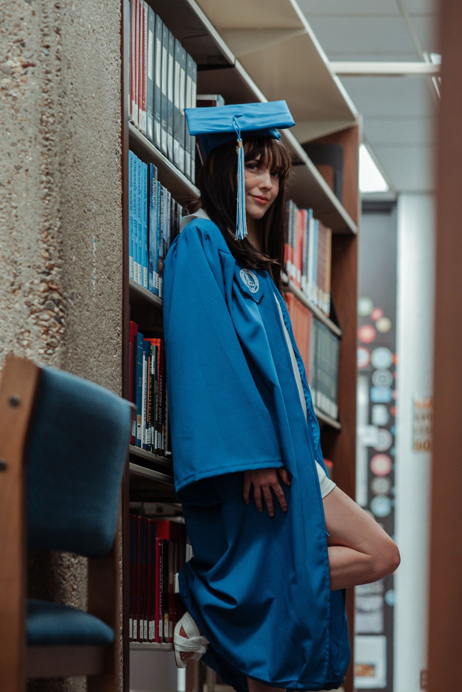 Female graduate in blue cap and gown leaning against library bookshelf smiling at camera.