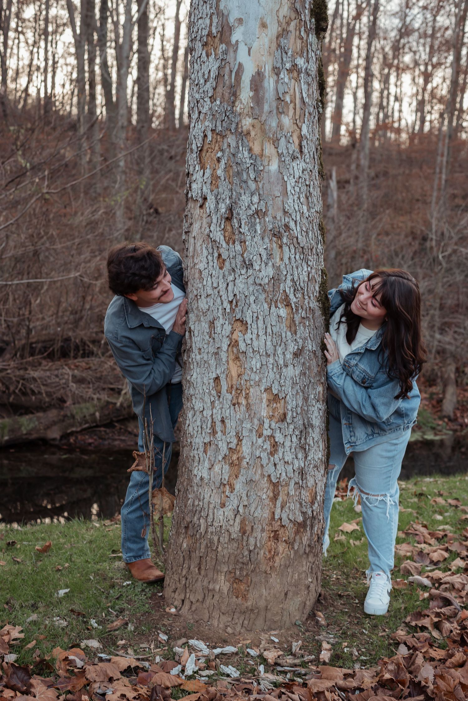 A playful couple laughs while hiding behind a large tree in a fall woodland setting.