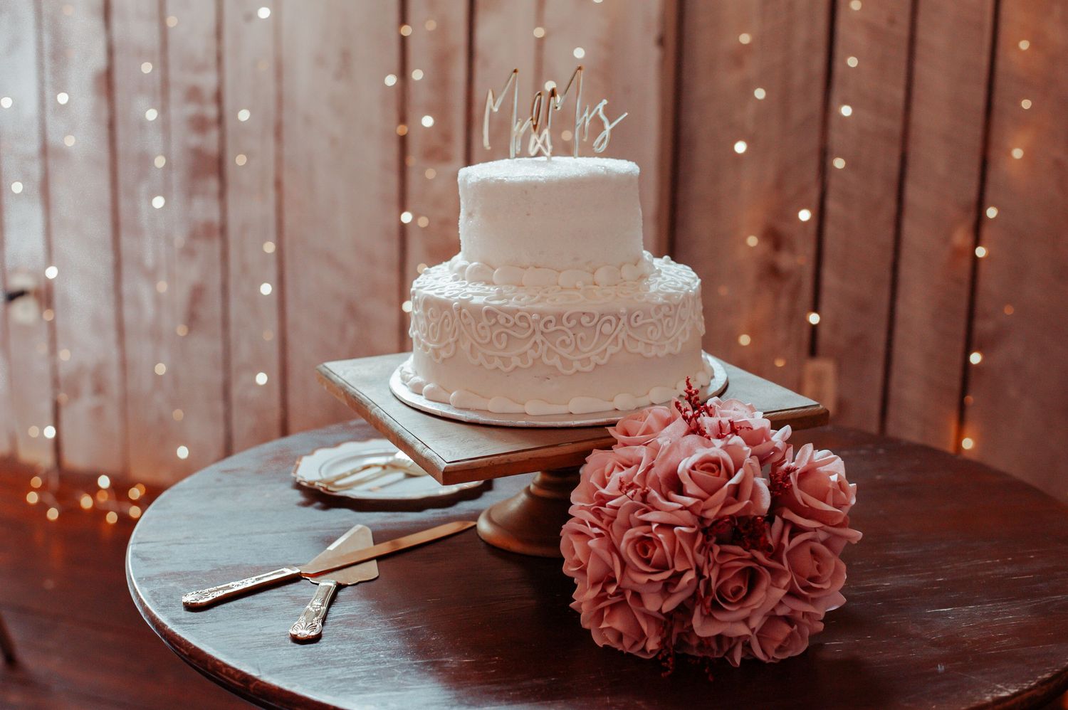 Two-tier white wedding cake with pink rose bouquet on wooden table against a bokeh light backdrop.