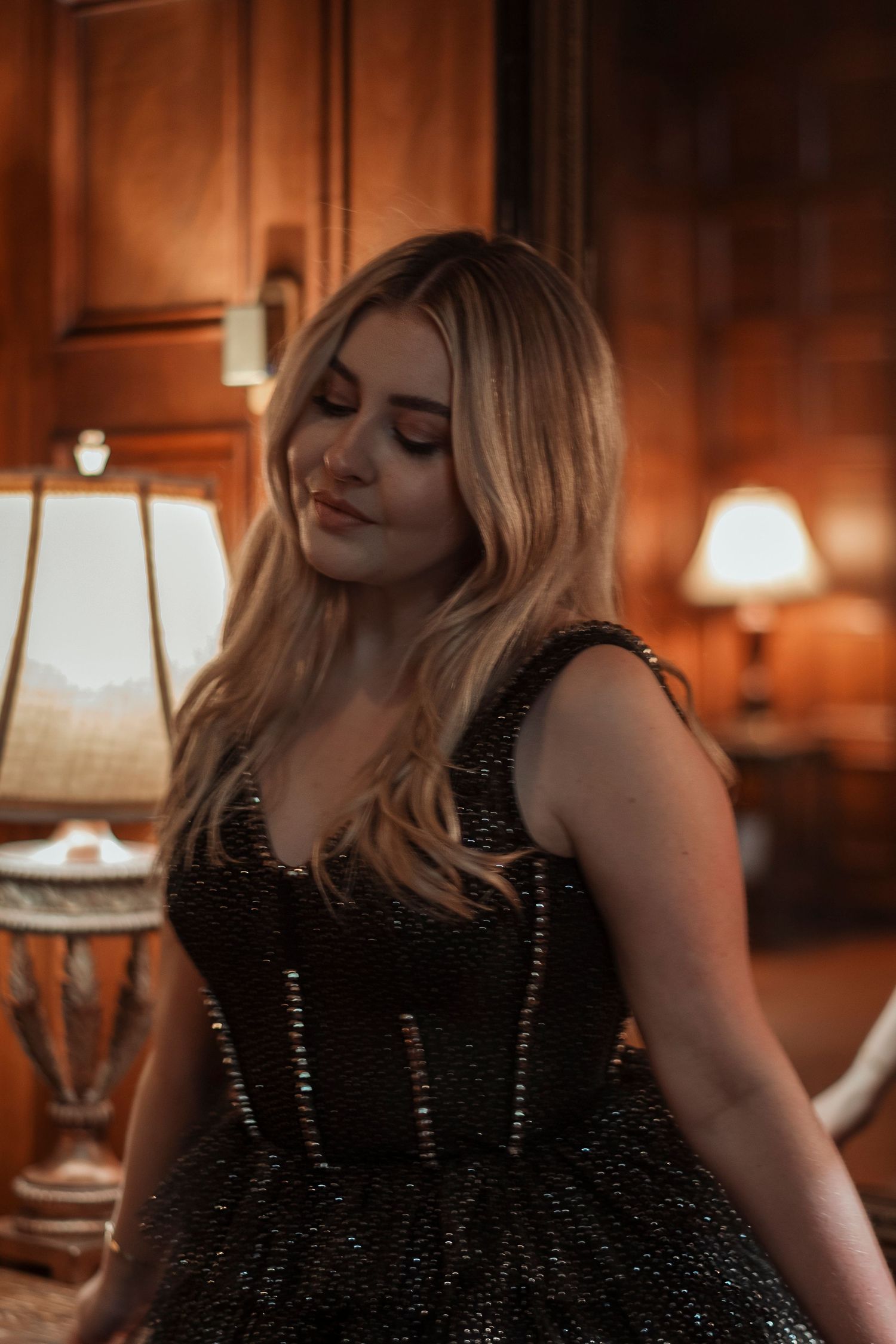 Blonde woman in black beaded corset dress posing in warmly lit wood-paneled elegant room.