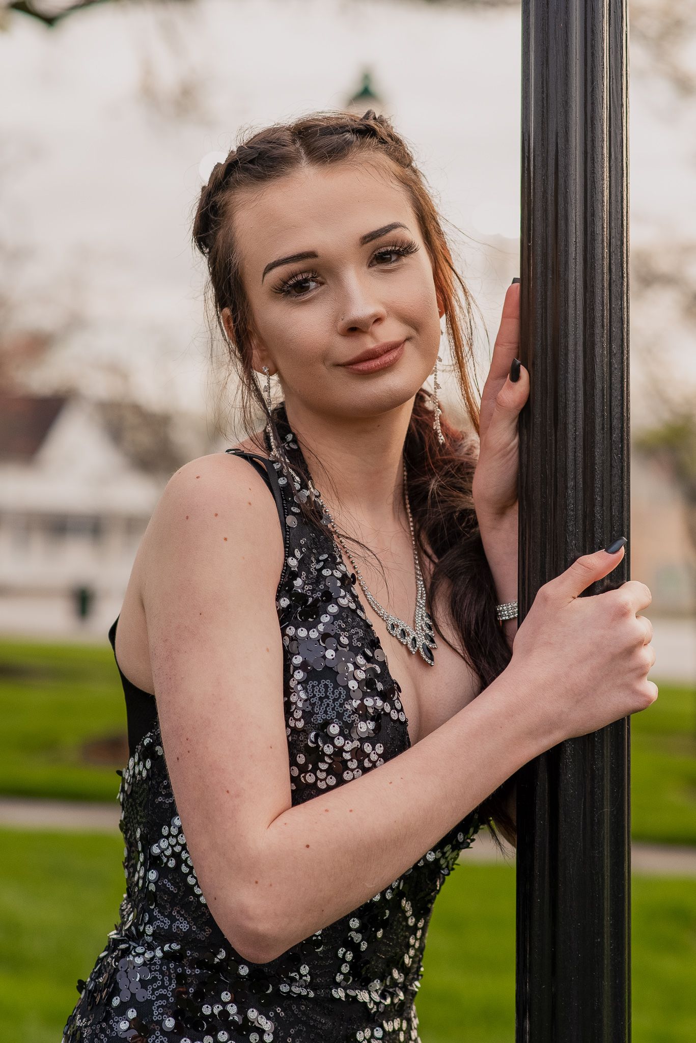 Young woman in black sequin dress leaning against a dark pole in an outdoor park setting.