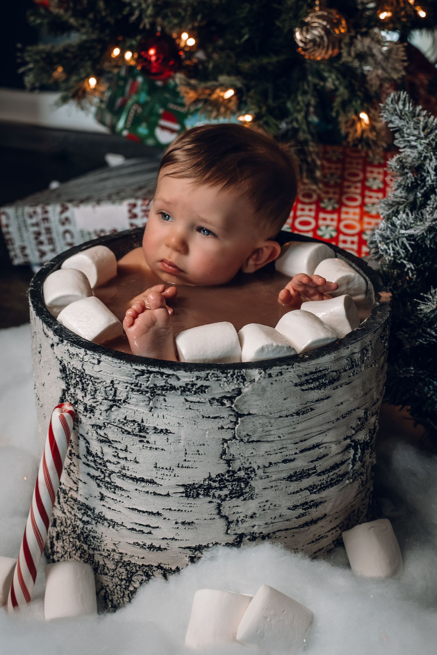 Cute baby rests chin on marshmallow-filled birch barrel beside Christmas tree with candy cane.