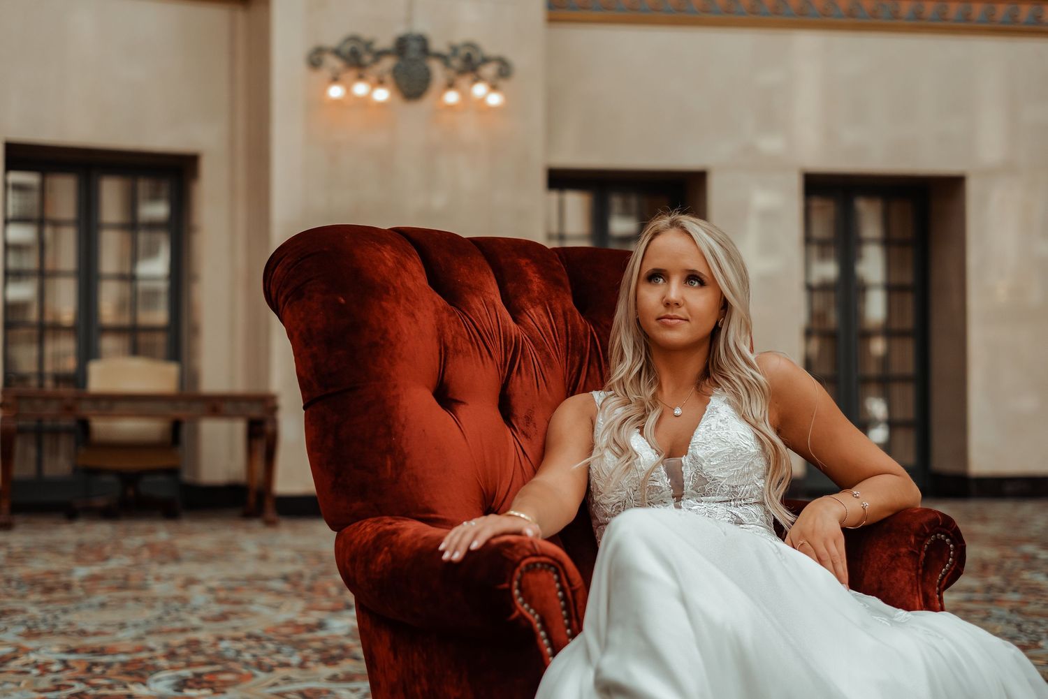 Bride in white gown seated elegantly in a red velvet armchair inside a grand vintage venue.