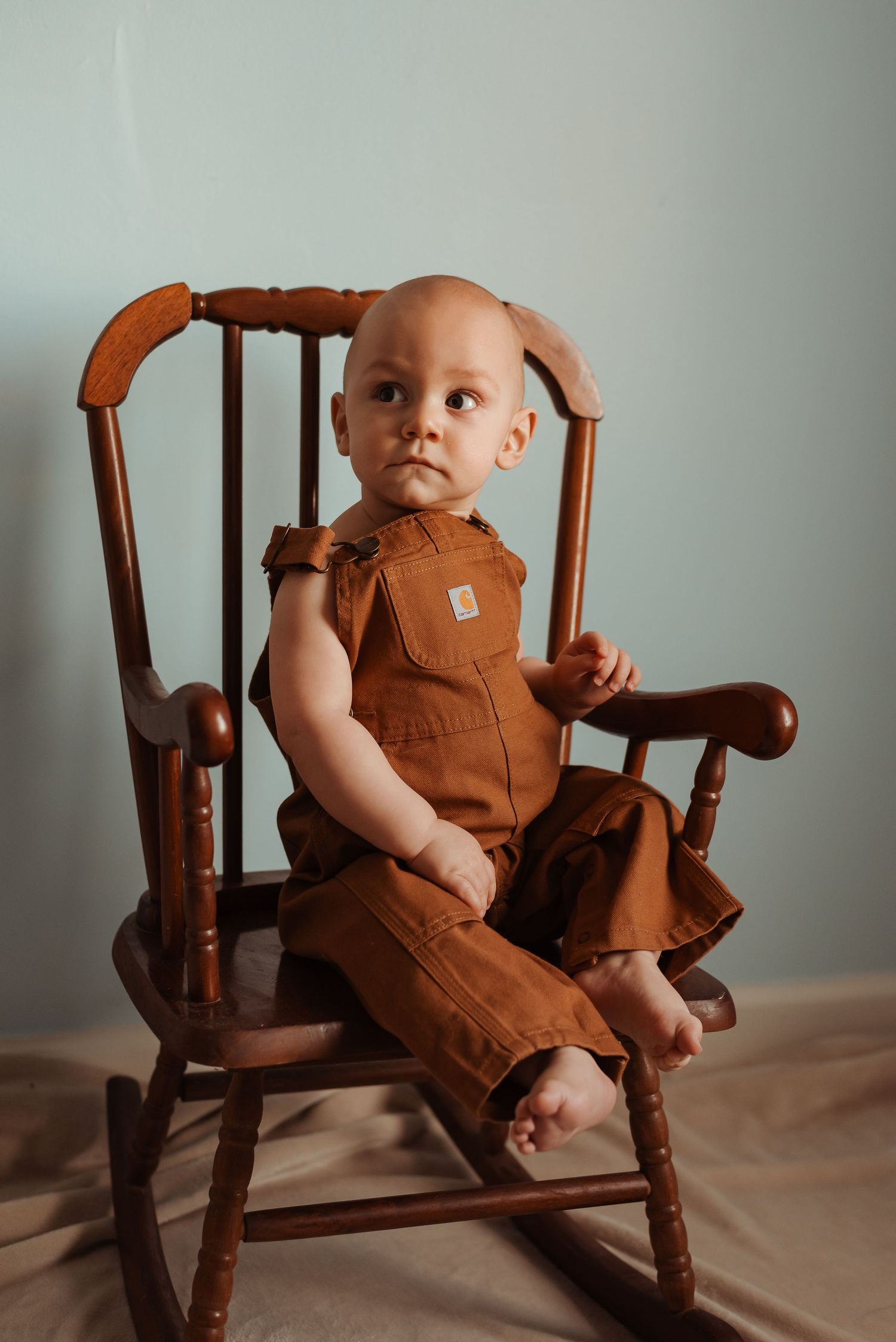 Baby in brown Carhartt overalls looks up while seated in a classic wooden rocking chair.