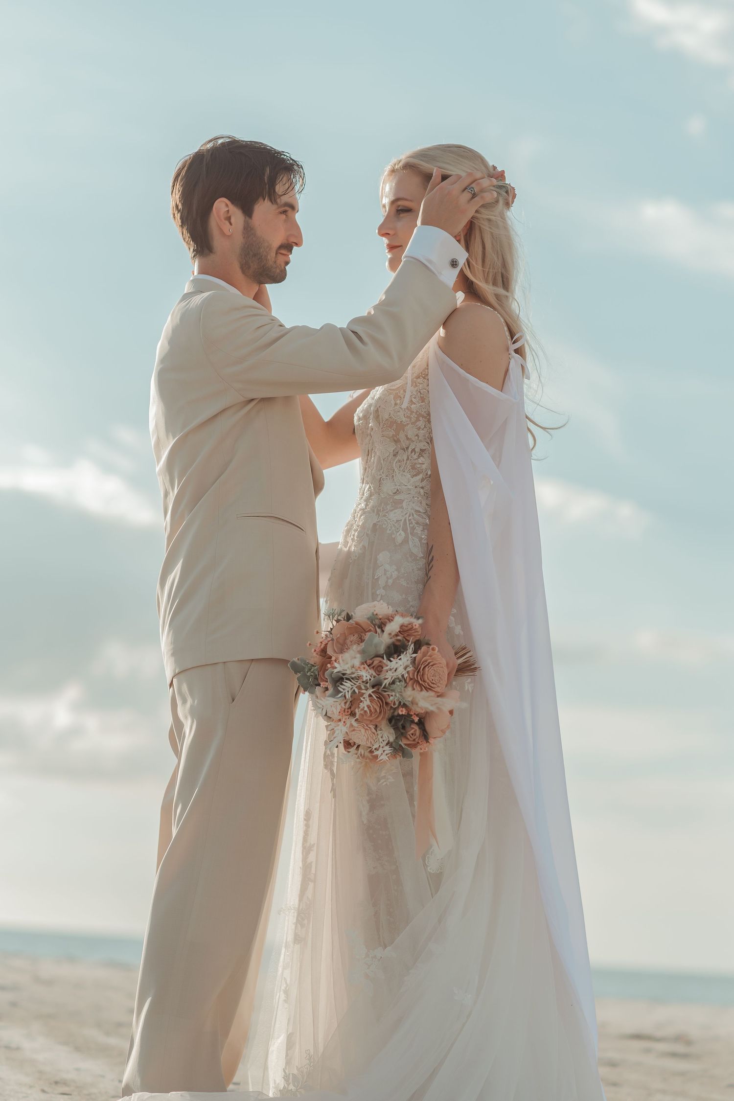 Groom tenderly touching bride's face as they stand together on a sunny beach wedding day.