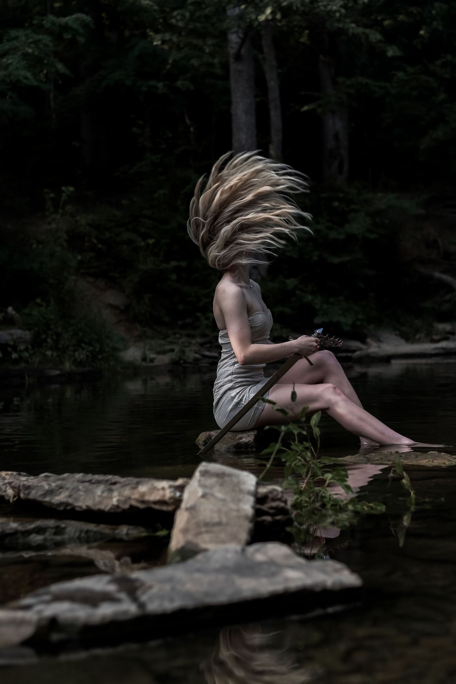 Woman in gray dress sits on rocks by water with hair dramatically flying over her face.