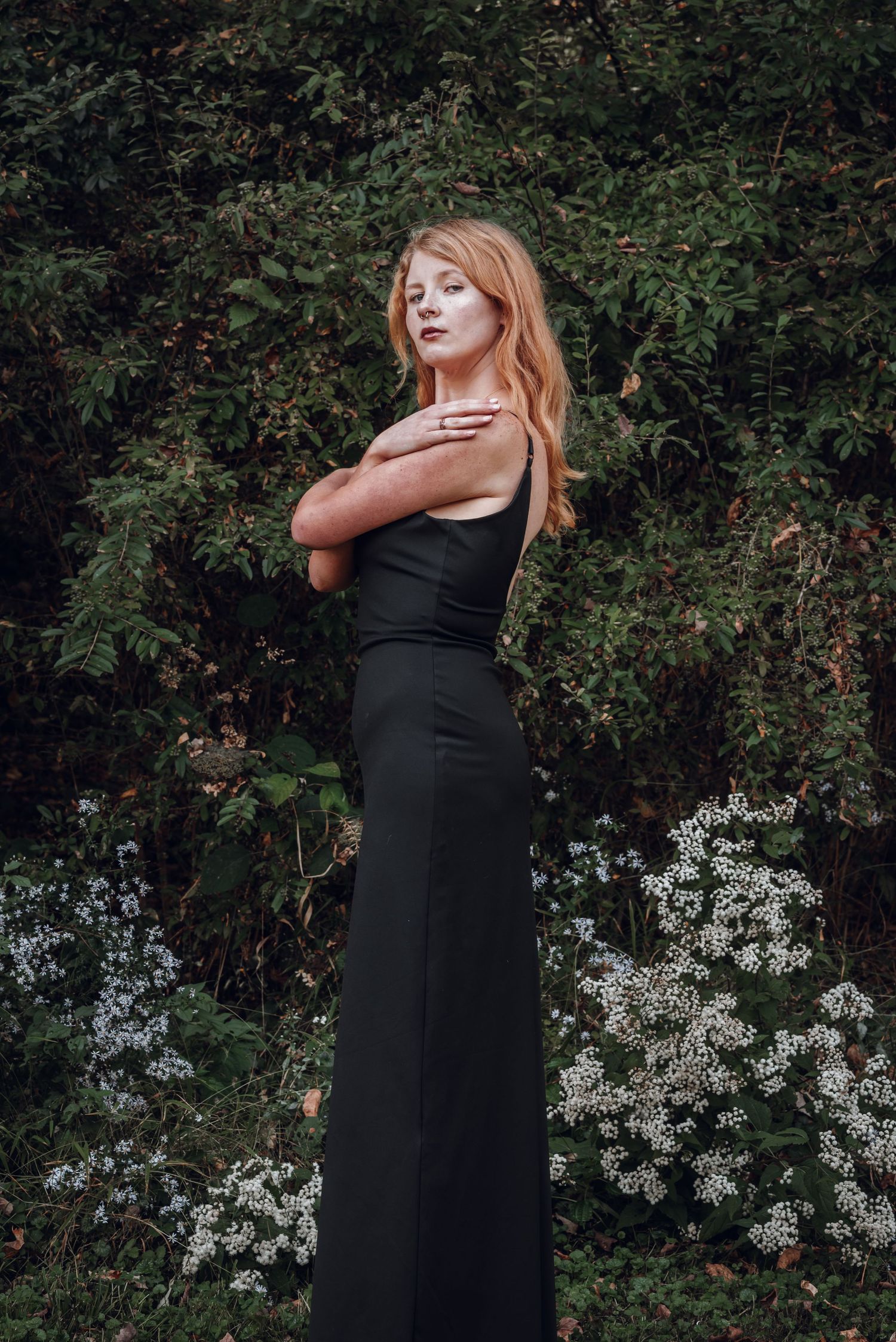 Young woman in elegant black gown with arms crossed, posing before green bushes and white flowers.