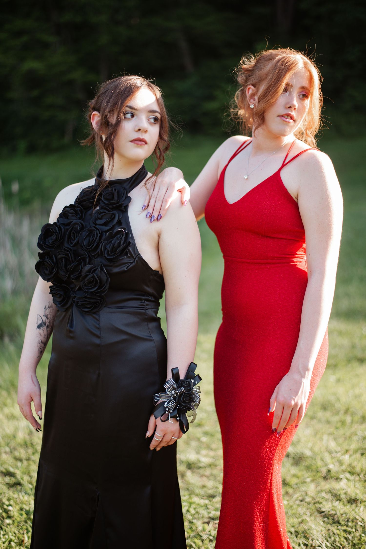 Two prom girls stand in field, one in black satin gown and one in red fitted dress.