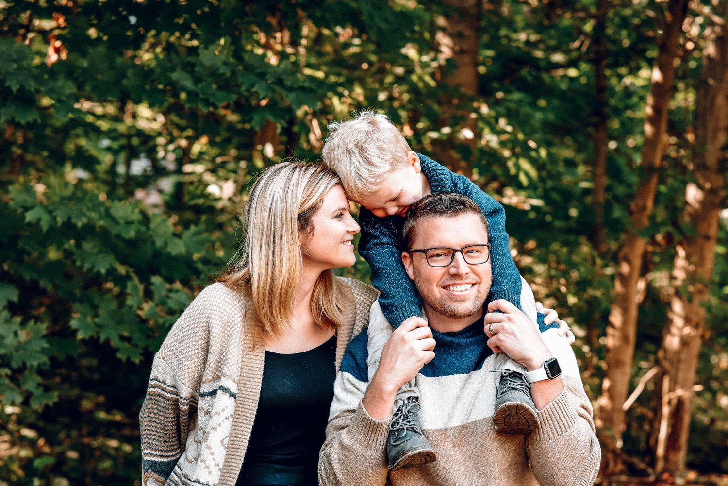 Smiling couple with young child on fathers shoulders poses outdoors among green trees for family photos.