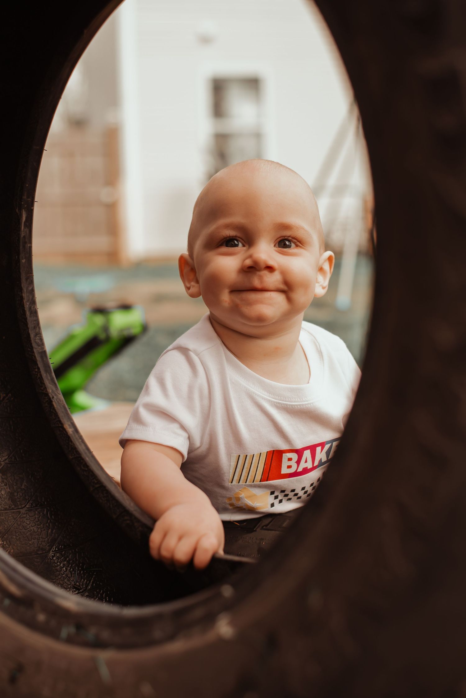 Smiling baby boy peers through a round tire opening wearing a white racing-themed shirt.