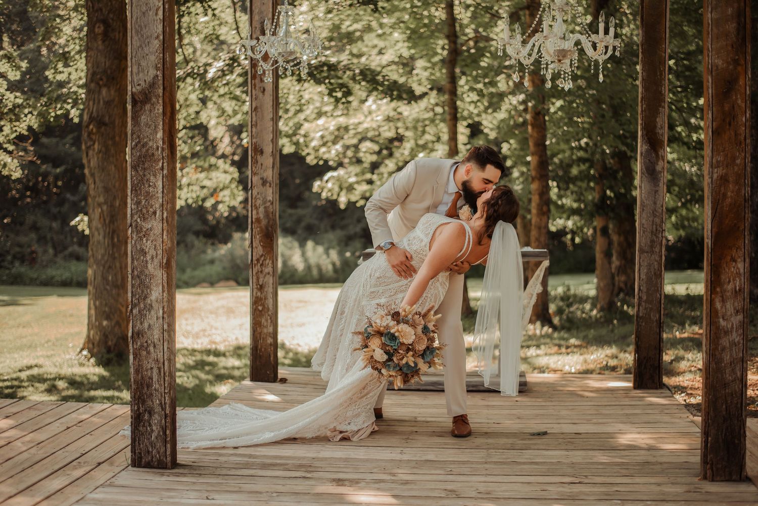 Bride and groom sharing romantic dip kiss on wooden deck surrounded by lush green trees.