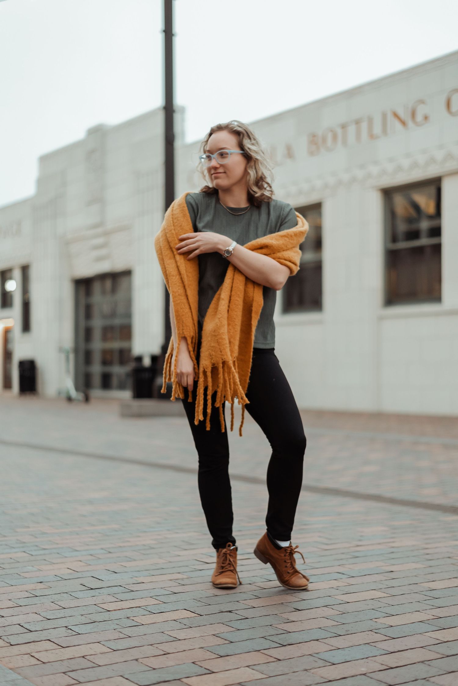 Young woman in olive tee, black leggings, and mustard fringe scarf posing on brick plaza downtown.