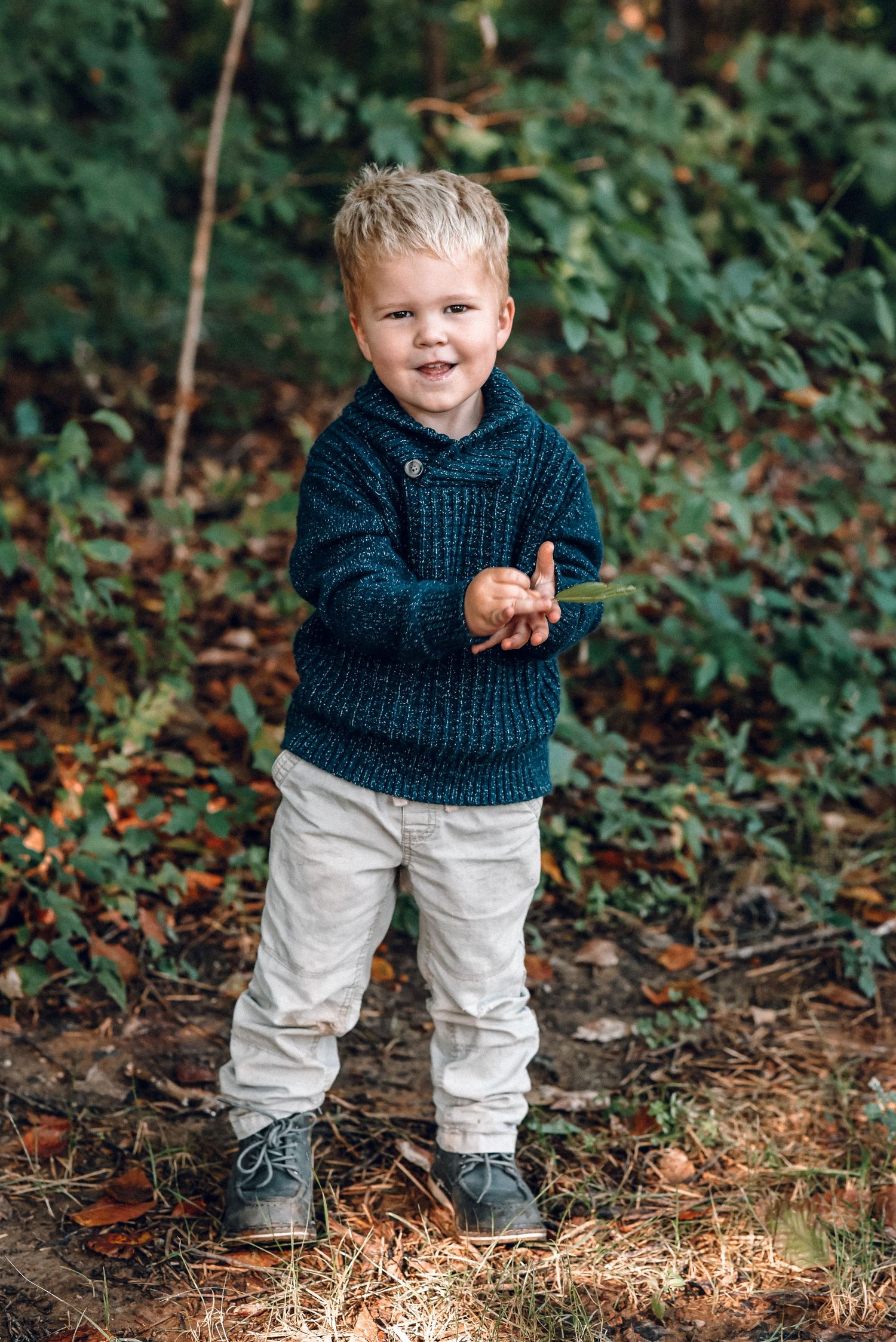 Smiling toddler boy in teal knit sweater stands outdoors among autumn foliage holding a leaf.