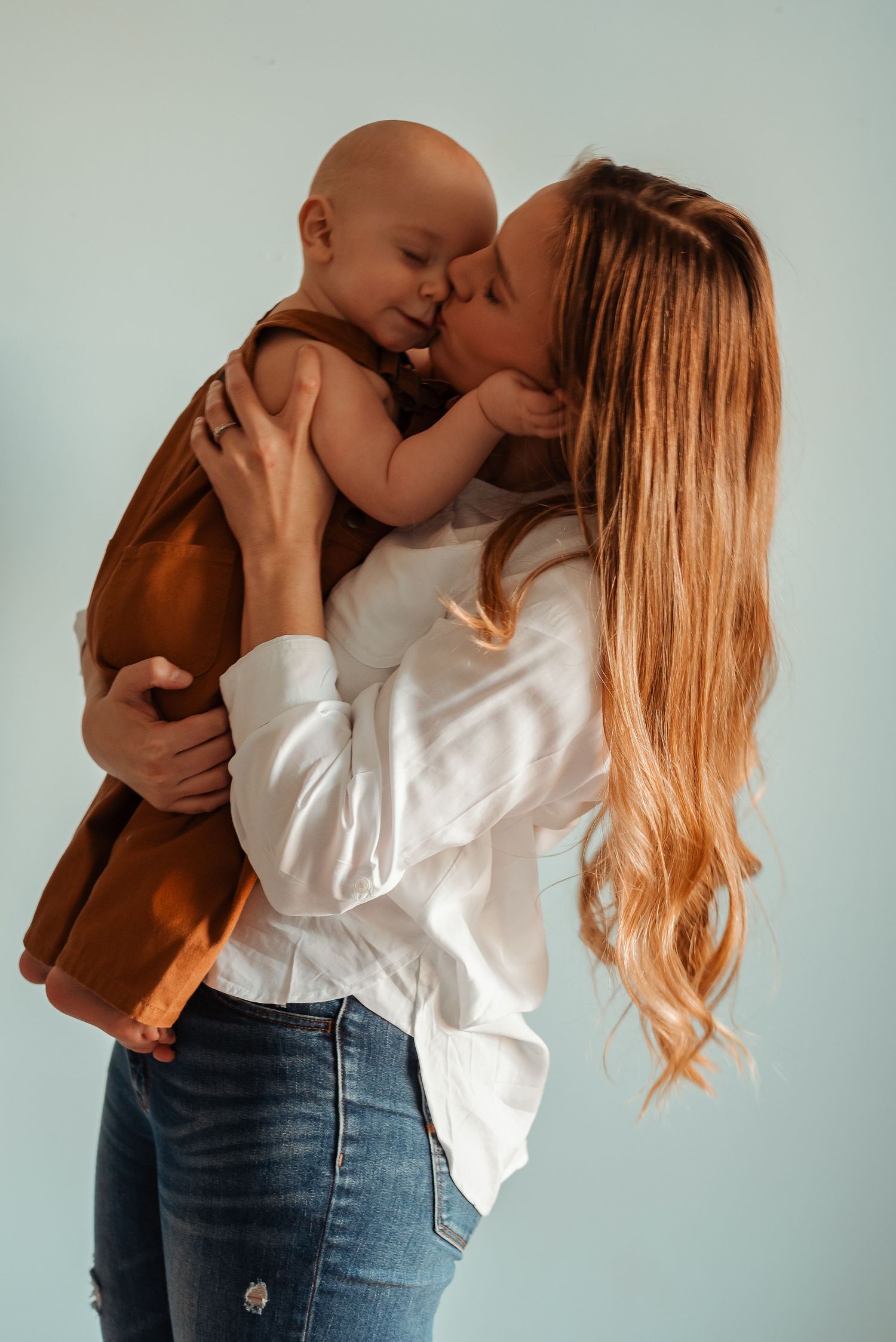 Loving mother with long blonde hair kisses her baby dressed in brown overalls.