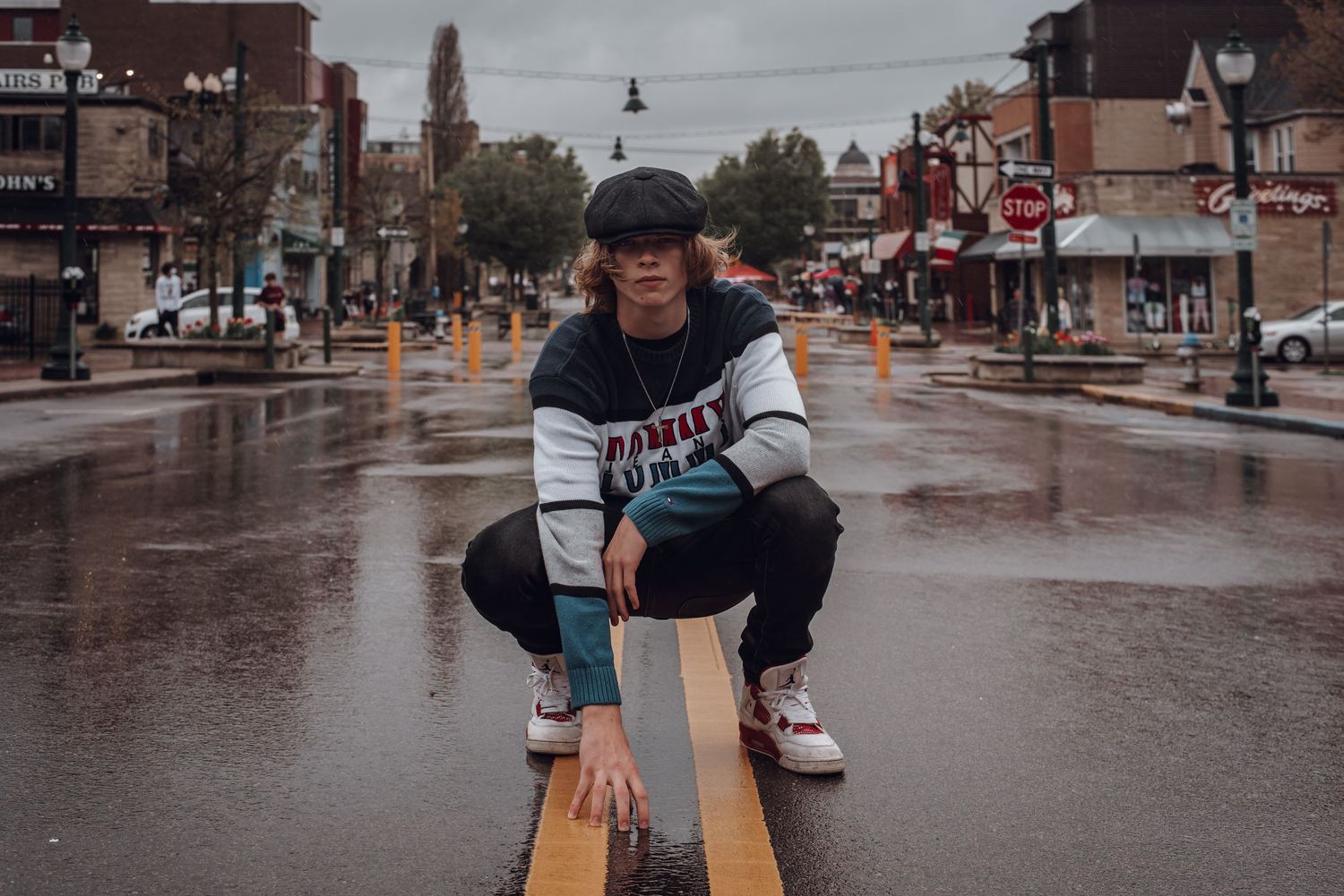 Young person crouching on wet city street in rainy urban setting wearing newsboy cap.