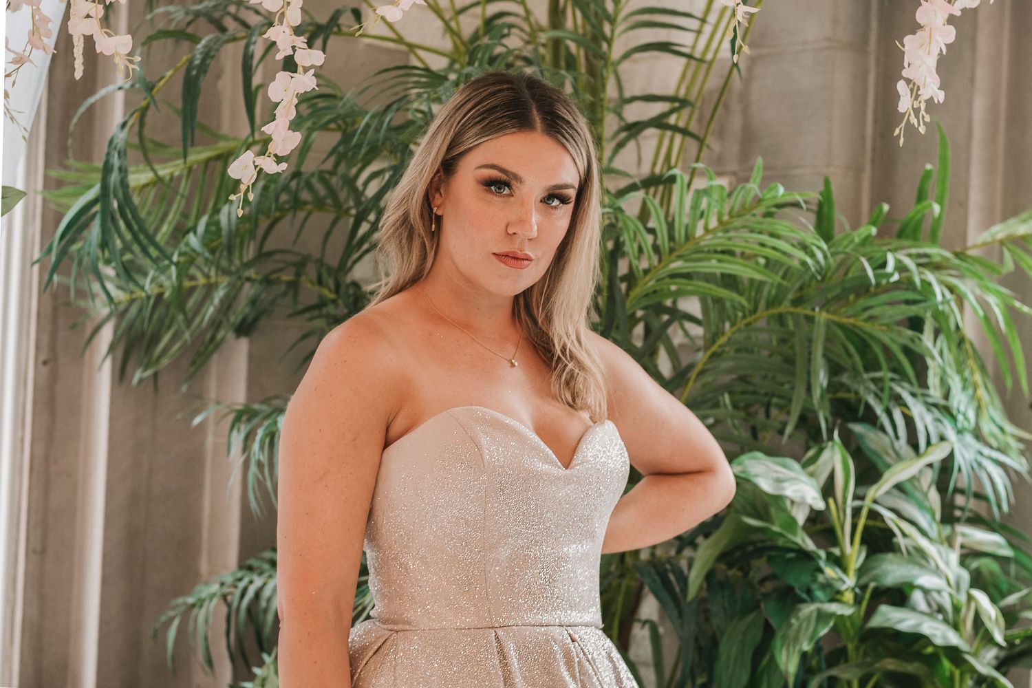 Woman in strapless rose gold ballgown posing seriously amid lush tropical greenery and white orchids.