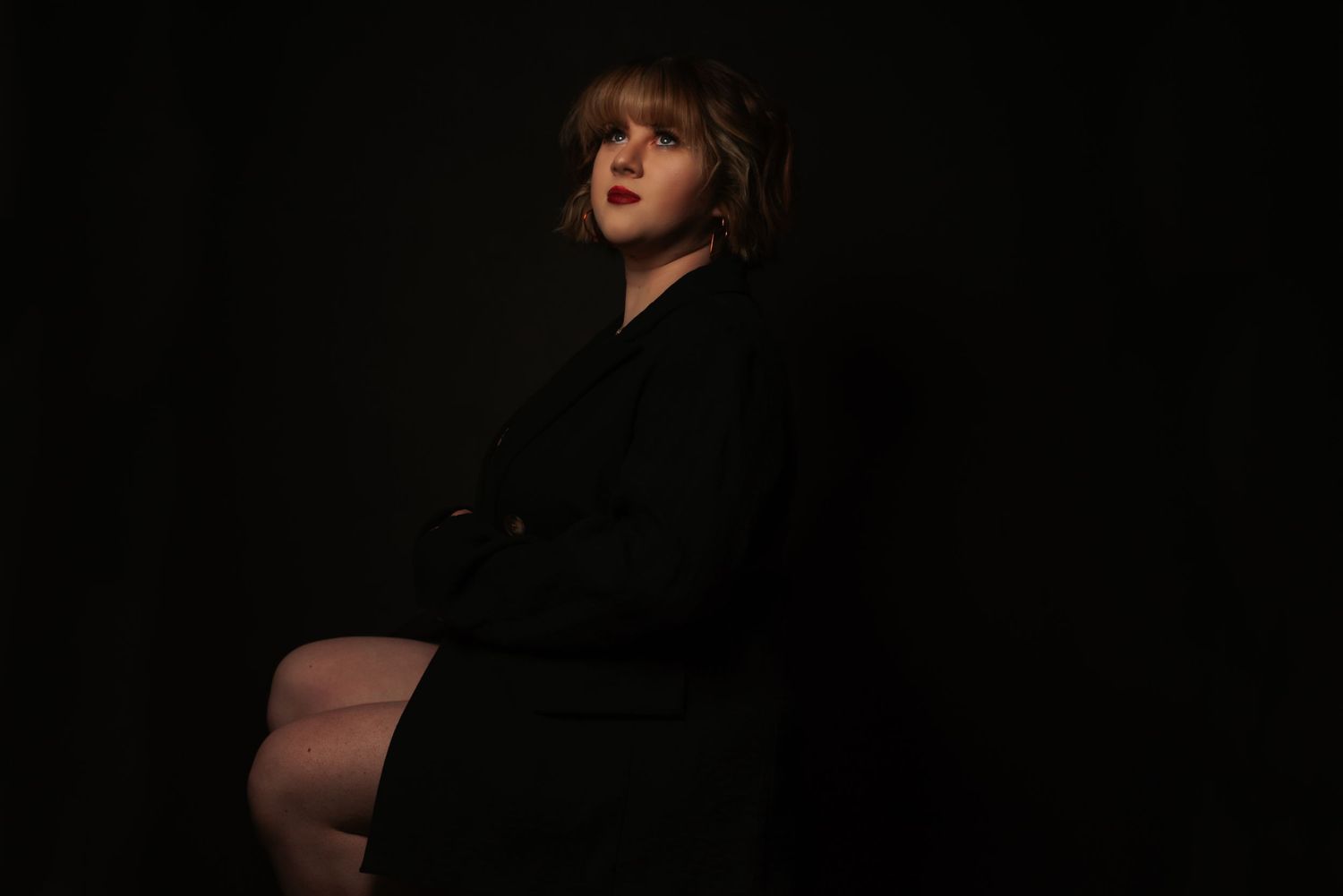 Woman in a black blazer seated against a dark background, gazing upward in a dramatic studio portrait.