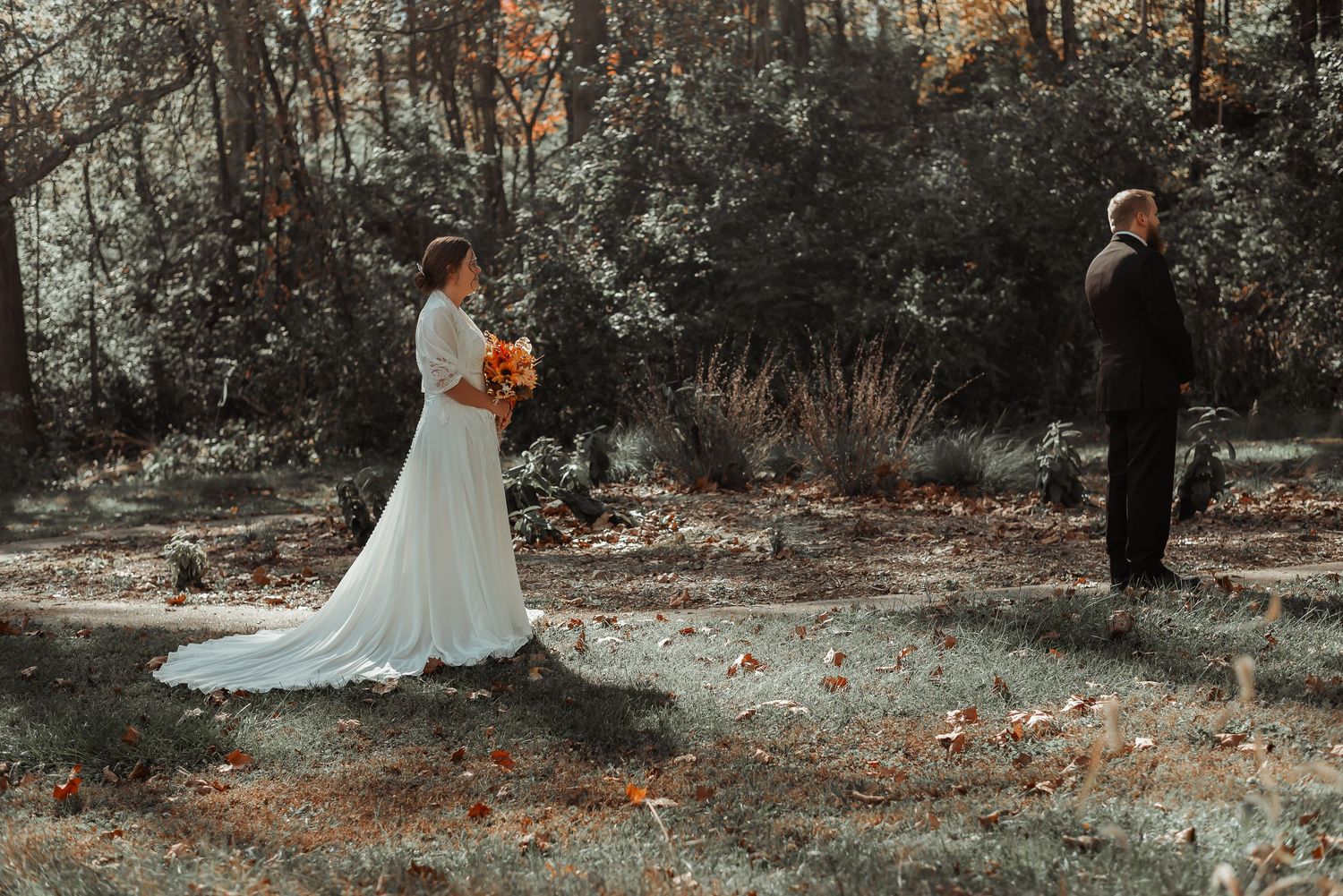 Bride holding orange bouquet stands behind groom in autumn forest.