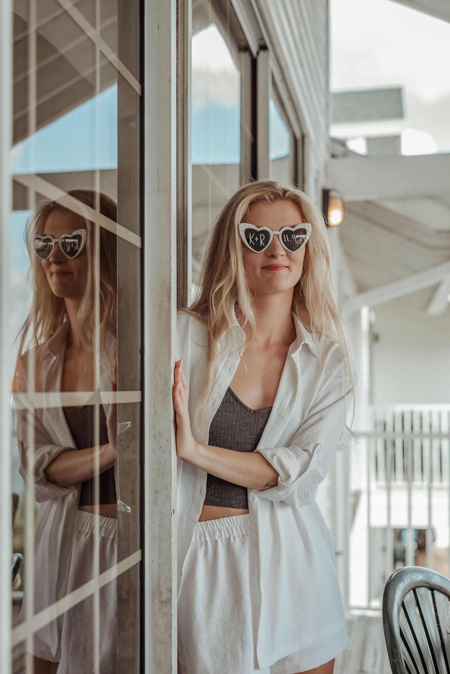 Blonde bride in white linen outfit wearing heart-shaped bride sunglasses leaning against glass door.