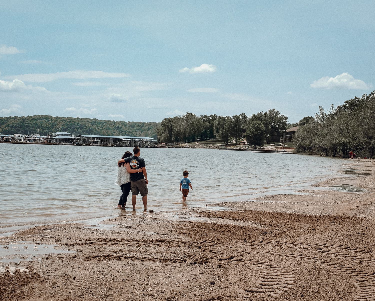 Family standing on sandy beach shore near calm lake waters under cloudy sky.