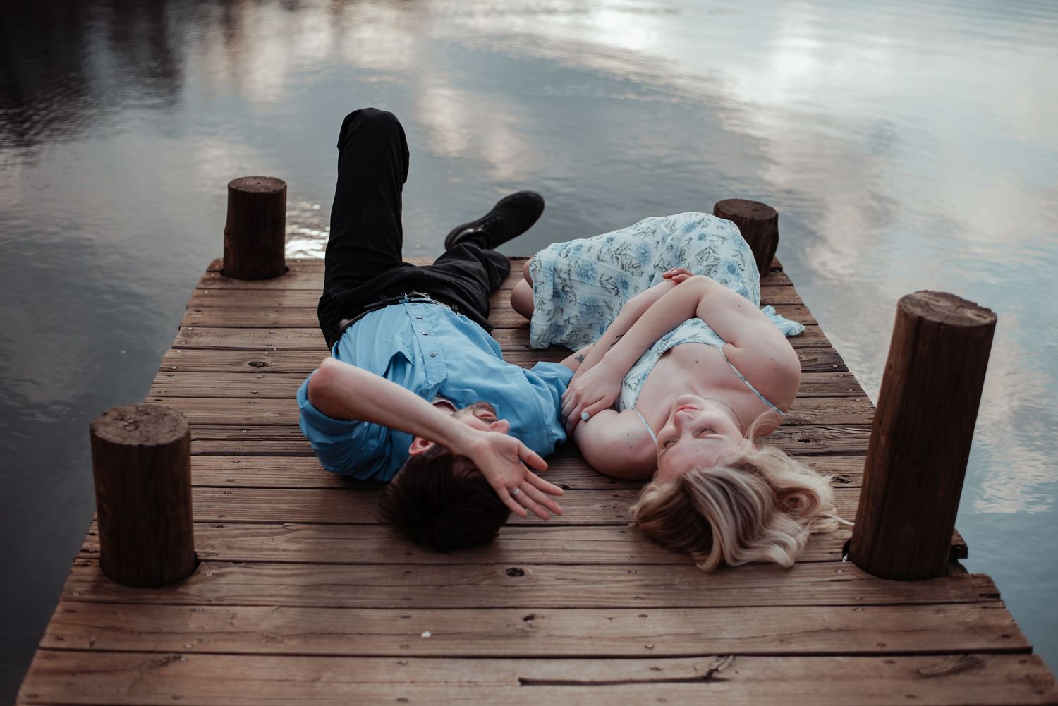 Romantic couple lying together on a wooden dock by a calm lake at dusk.