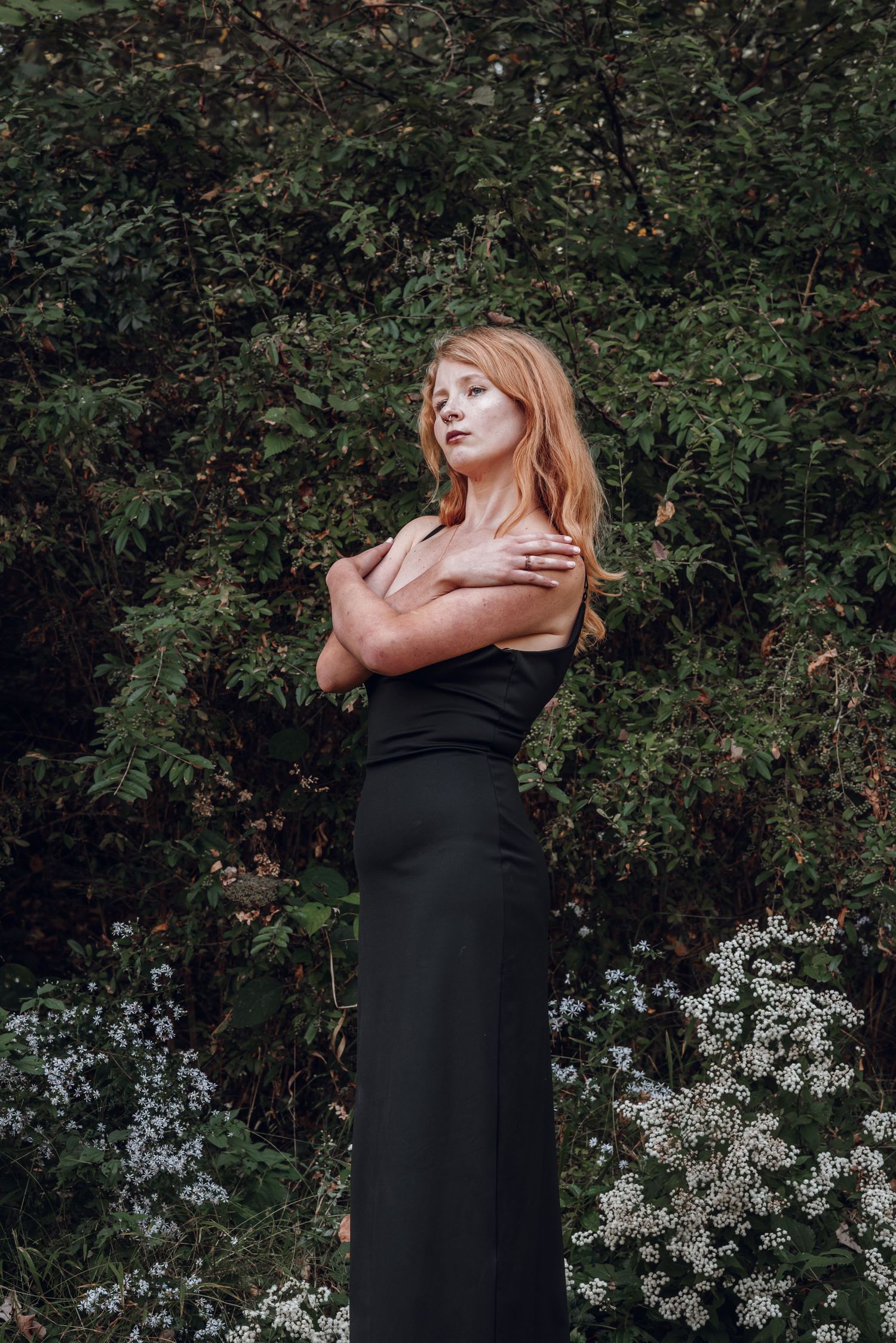 Elegant woman in black off-shoulder gown standing gracefully among green trees and white flowers.