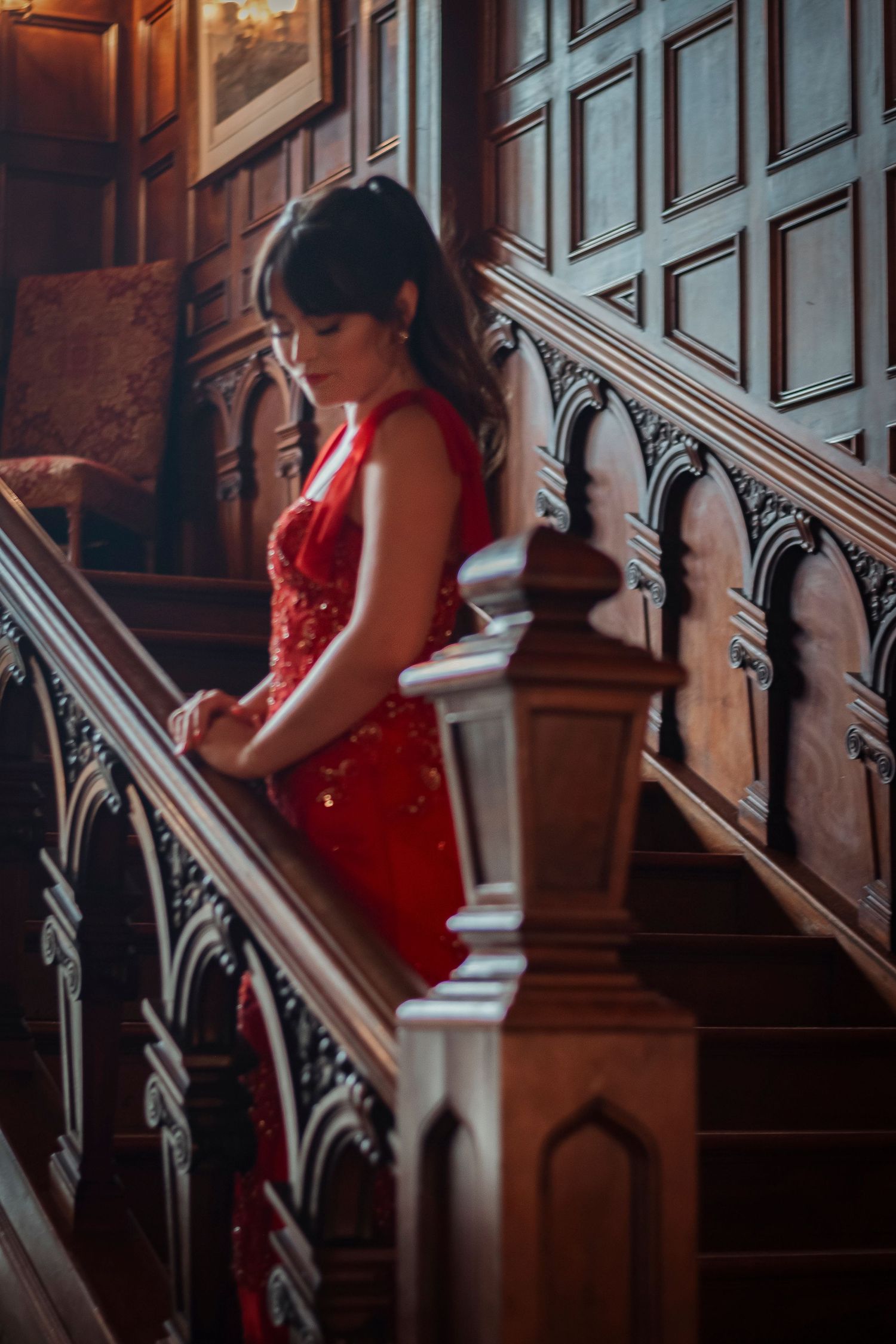 Woman in red sequin gown descending ornate wooden staircase in elegant historic mansion.