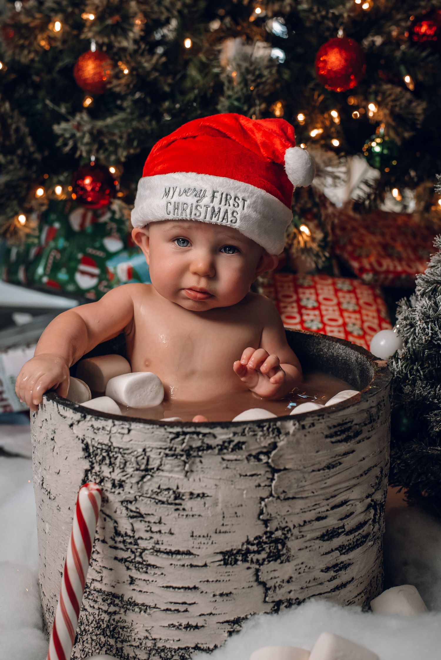 Adorable baby in Santa hat sits in rustic barrel with marshmallows for first Christmas photo.