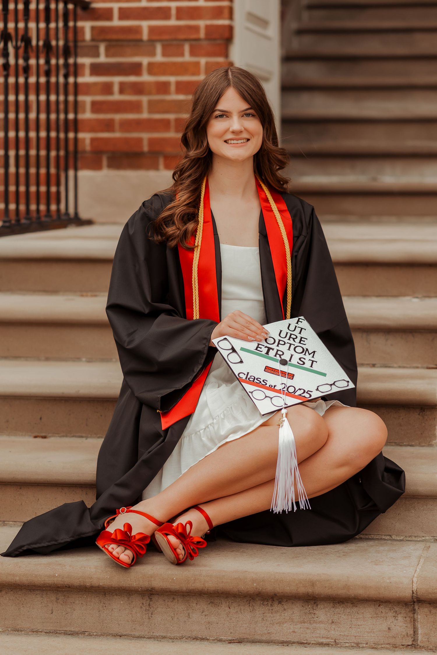 Smiling graduate in black gown with red stole sitting on steps, holding decorated cap for Class of 2025.